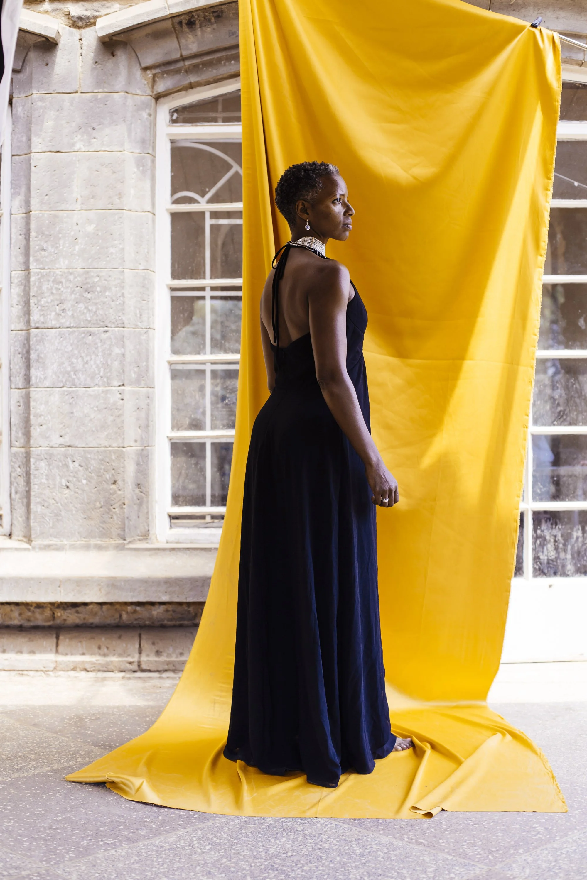 A woman in a long black dress stands barefoot on yellow fabric that extends from a backdrop to the floor, with a stone wall and large window in the background.