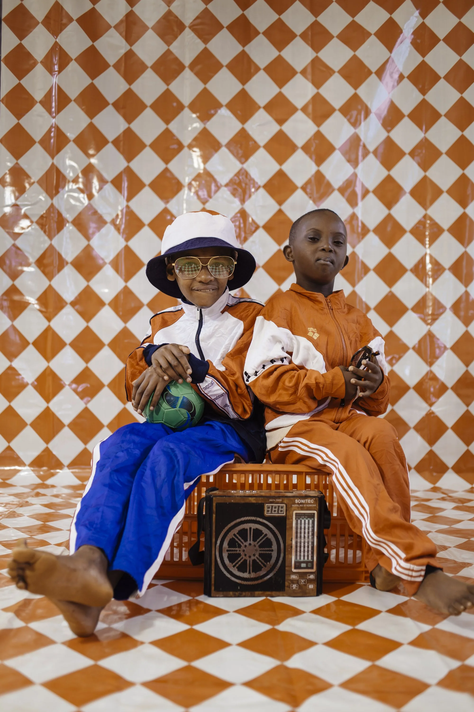 Two children dressed in orange and white tracksuits sitting indoors on a patterned orange and white checkered floor with a matching backdrop. One child is wearing sunglasses and a sunhat, holding a green soccer ball. The other child is holding a toy 