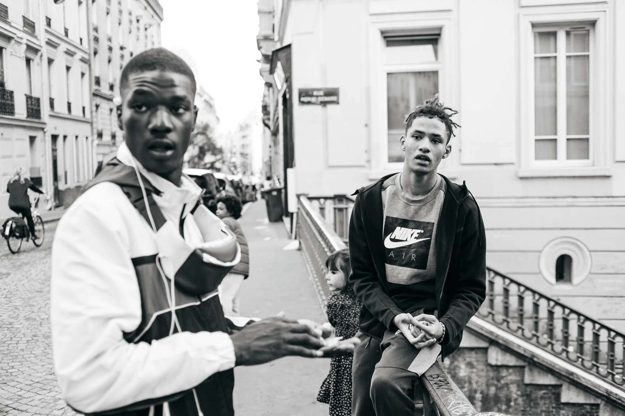 Black and white photo of two young men standing on a city sidewalk. The young man on the left is wearing a jacket with a hood and is looking at the camera. The young man on the right is sitting on a railing, wearing a Nike jacket, and looking at the 