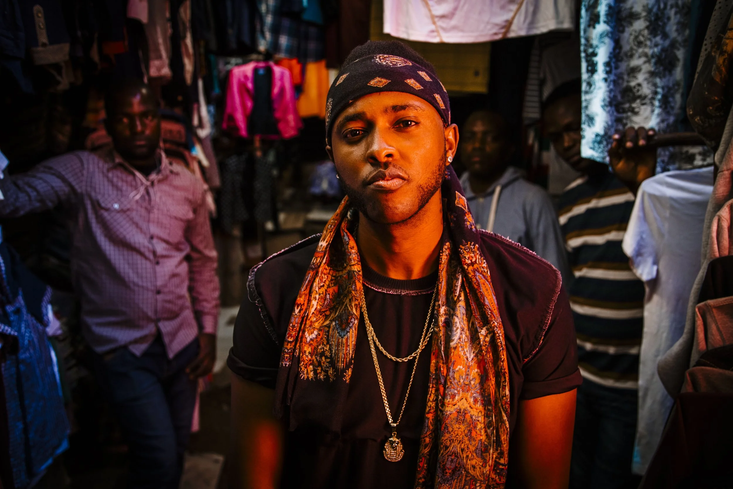 Young man with earrings, a patterned scarf, and jewelry, standing in a vibrant market or shop with people and colorful clothing in the background.