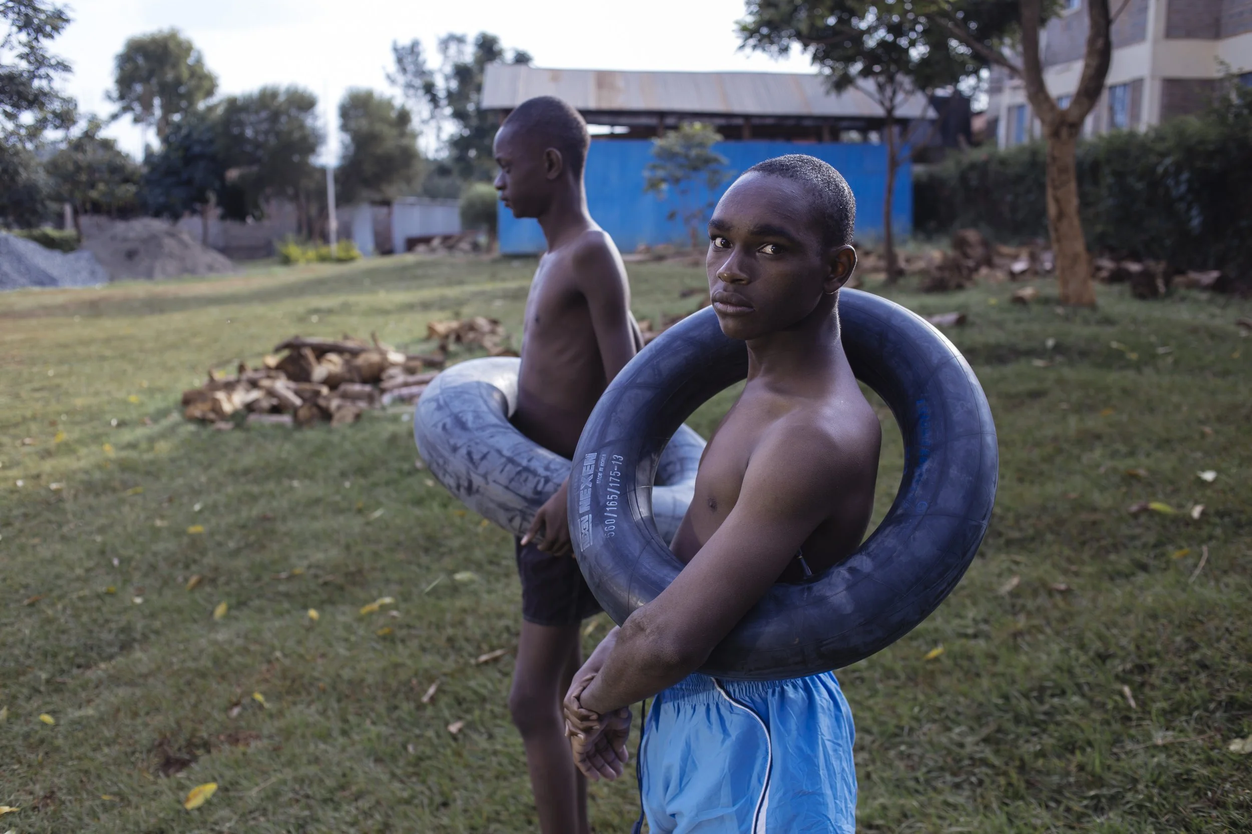 Two young boys without shirts, each holding inner tubes, standing outdoors on a grassy area with trees, a blue building, and stones in the background.