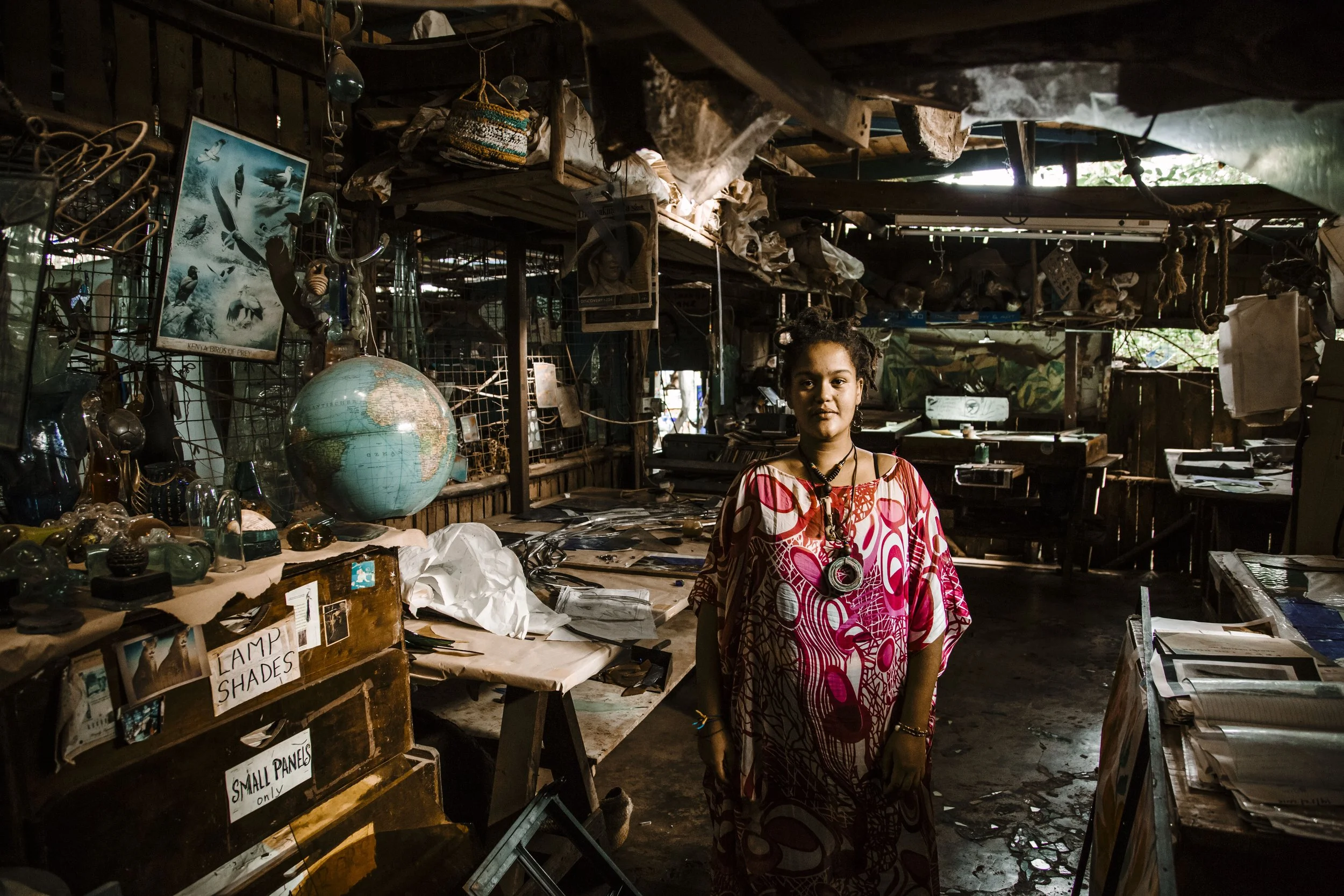 A young girl stands inside a cluttered store filled with various items like a globe, framed pictures, jewelry, and signs. The store appears to be made of wood with an assortment of objects on shelves and tables around her.