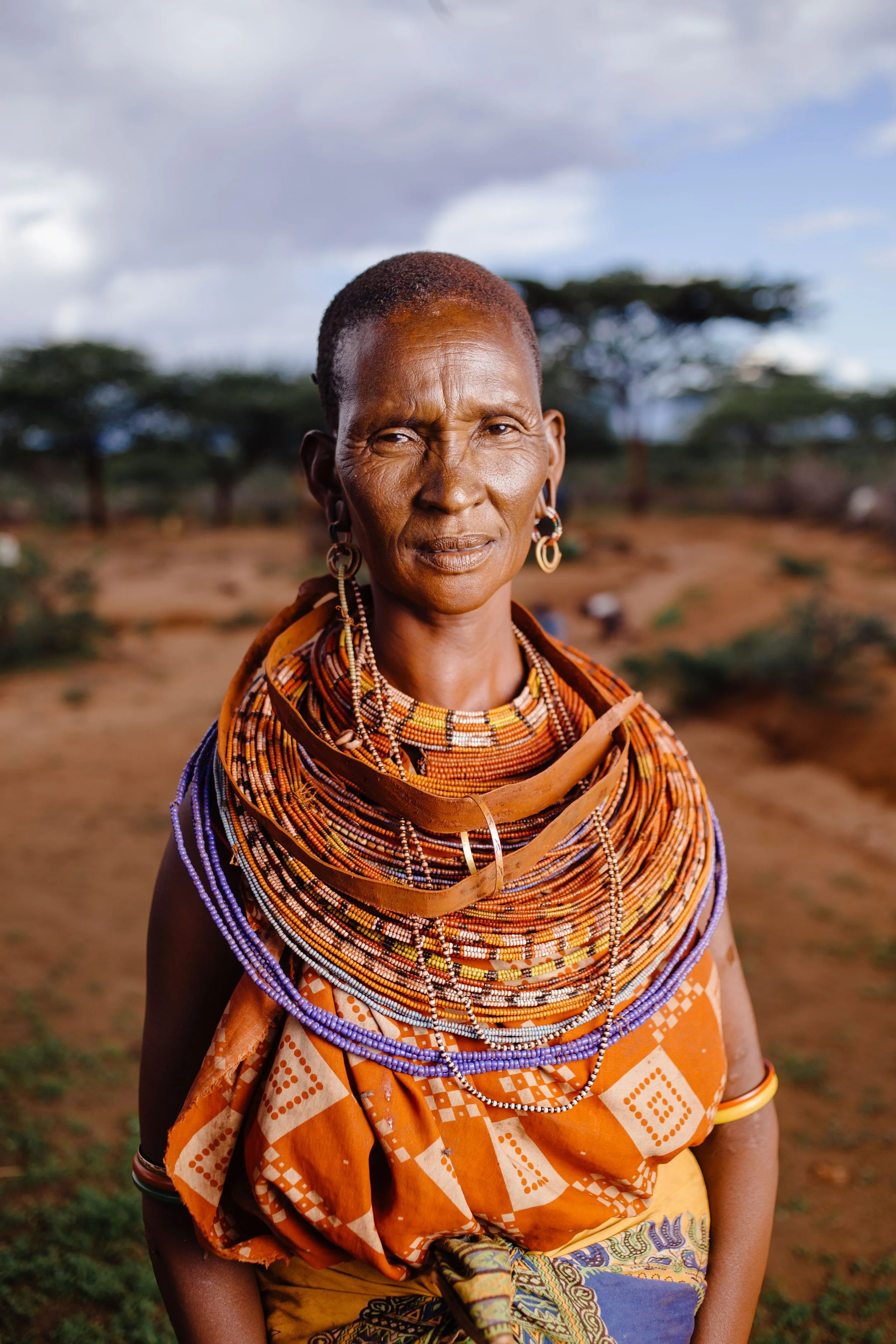 A woman from an African tribe, wearing traditional clothing and colorful bead jewelry, standing outdoors in a rural area with trees and cloudy sky in the background.
