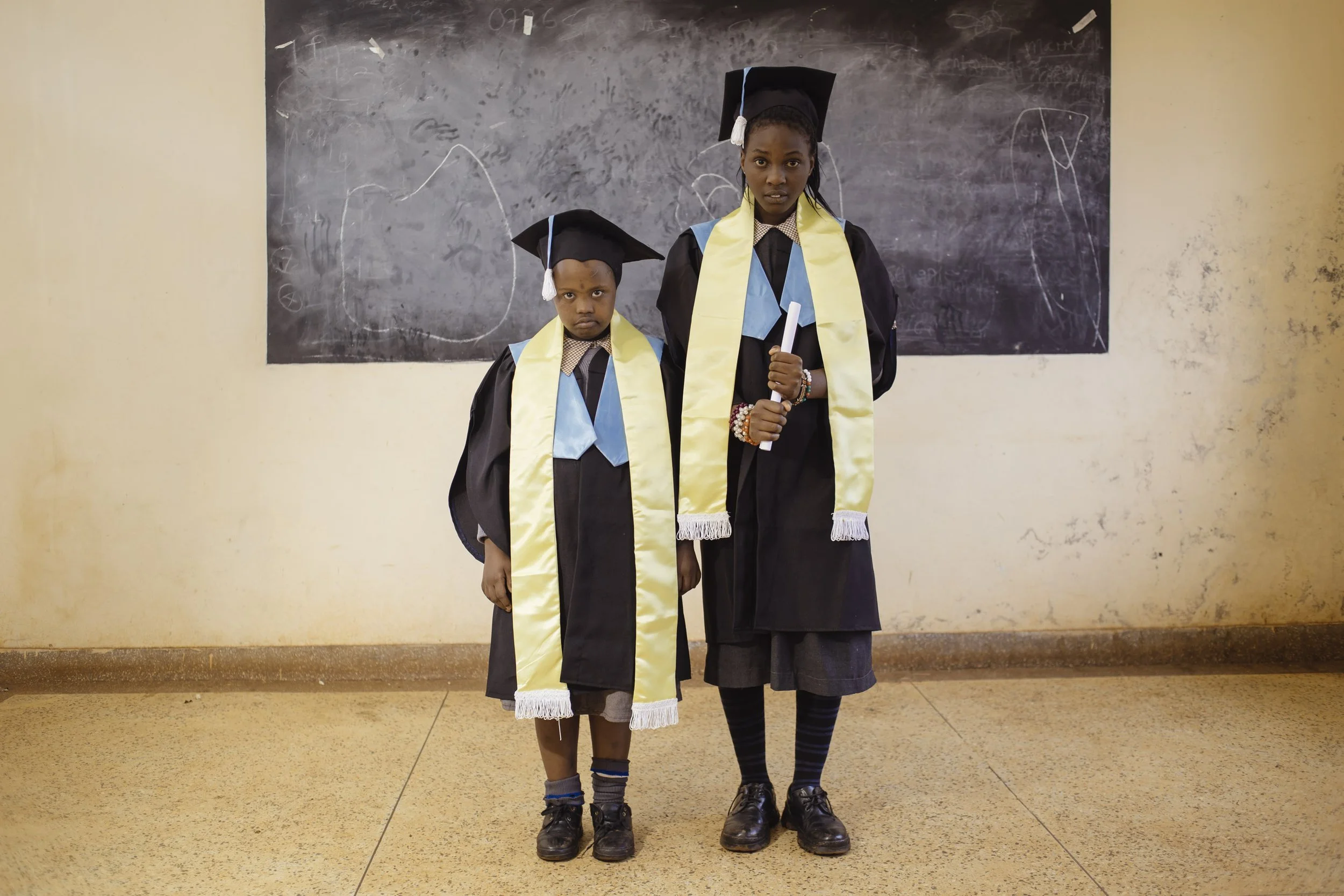 Two children in graduation caps and gowns stand in front of a blackboard. The older girl holds a rolled diploma, and both wear yellow sashes with blue accents.
