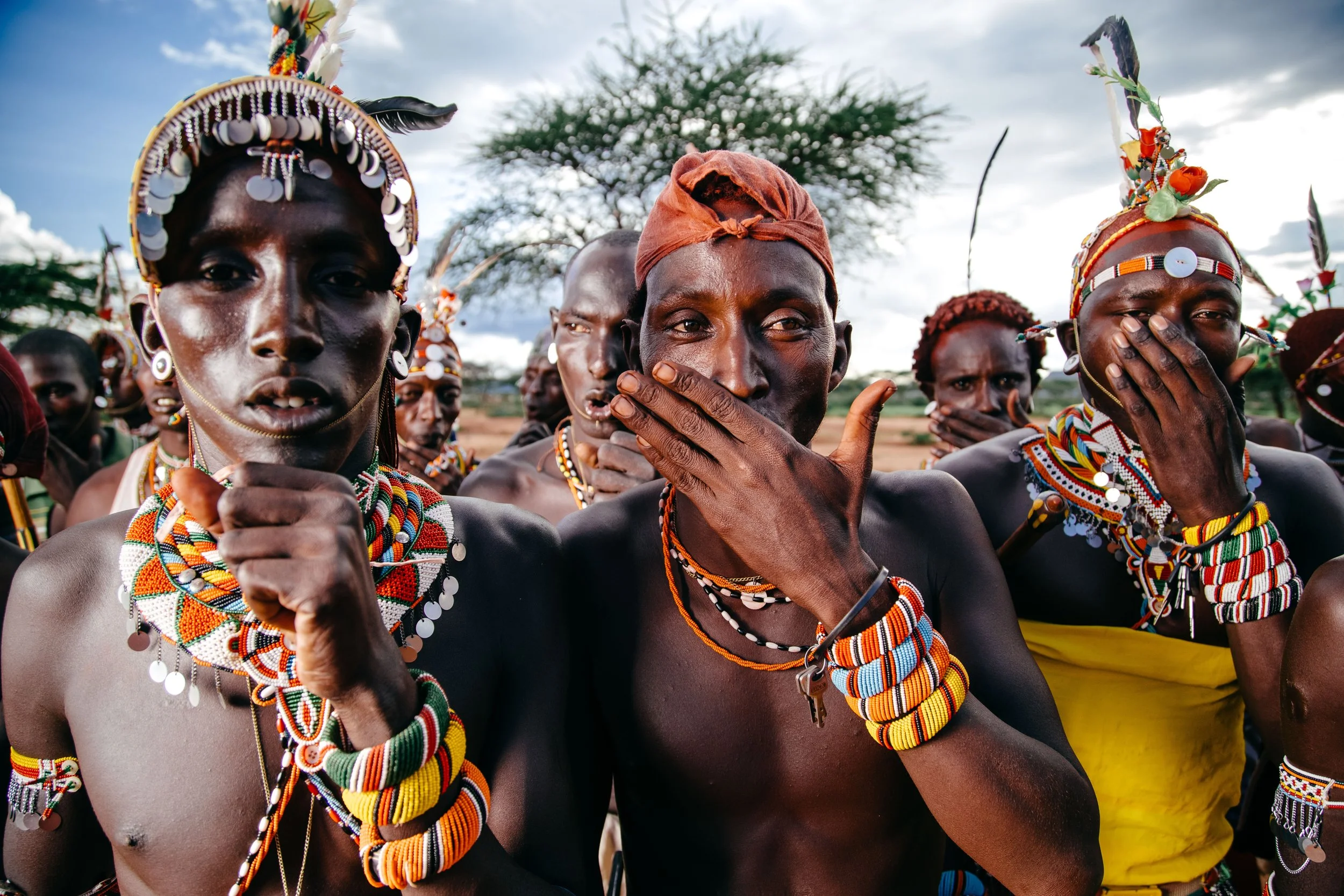 A group of Maasai men dressed in traditional bead jewelry and clothing, some covering their mouths with their hands, outdoors with cloudy sky and a tree in the background.