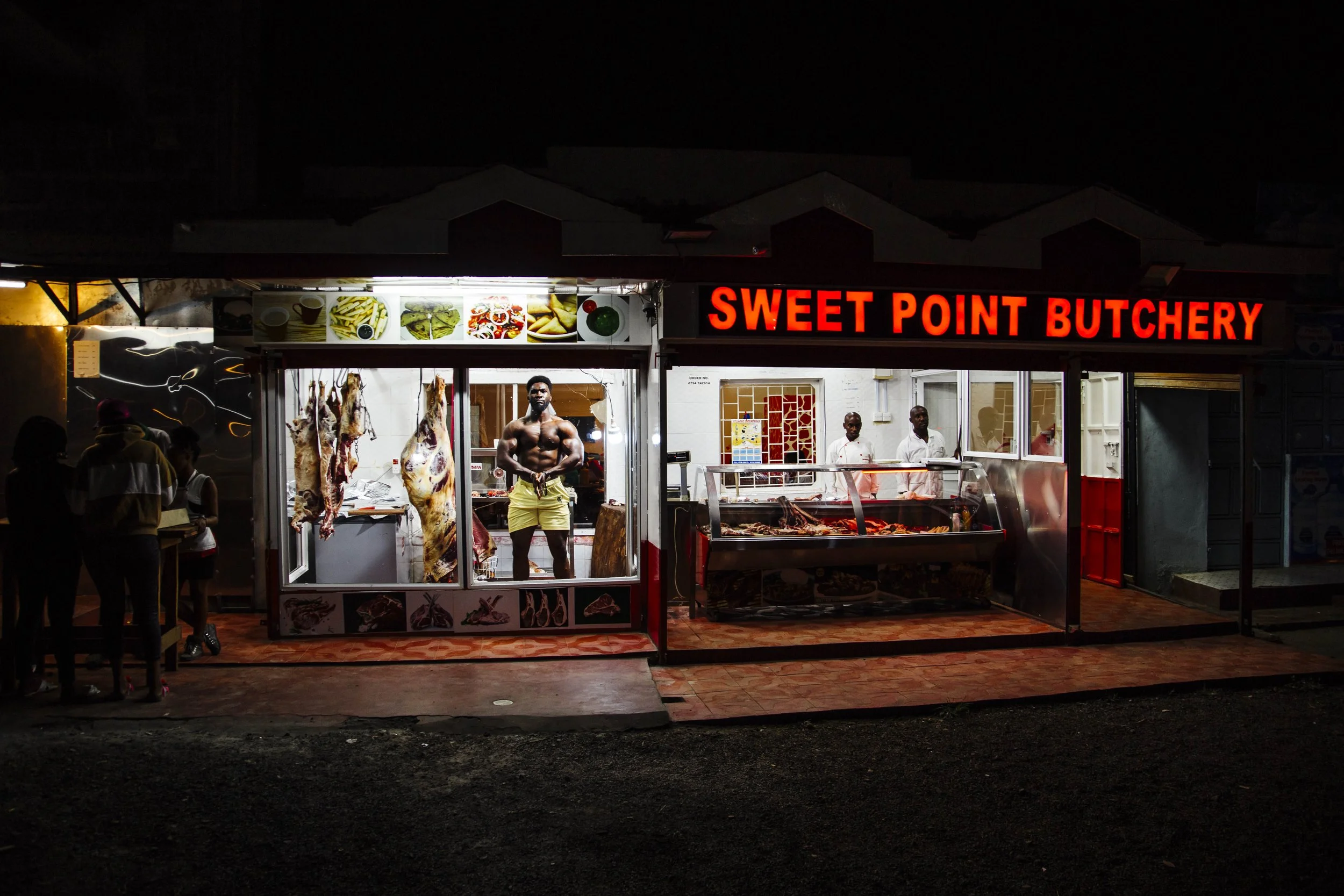 Nighttime view of a butchery shop named 'Sweet Point Butchery' with hanging meat, a muscular man in yellow shorts inside, and a display of meat cuts in the glass case. Two workers are behind the counter, and customers are outside.