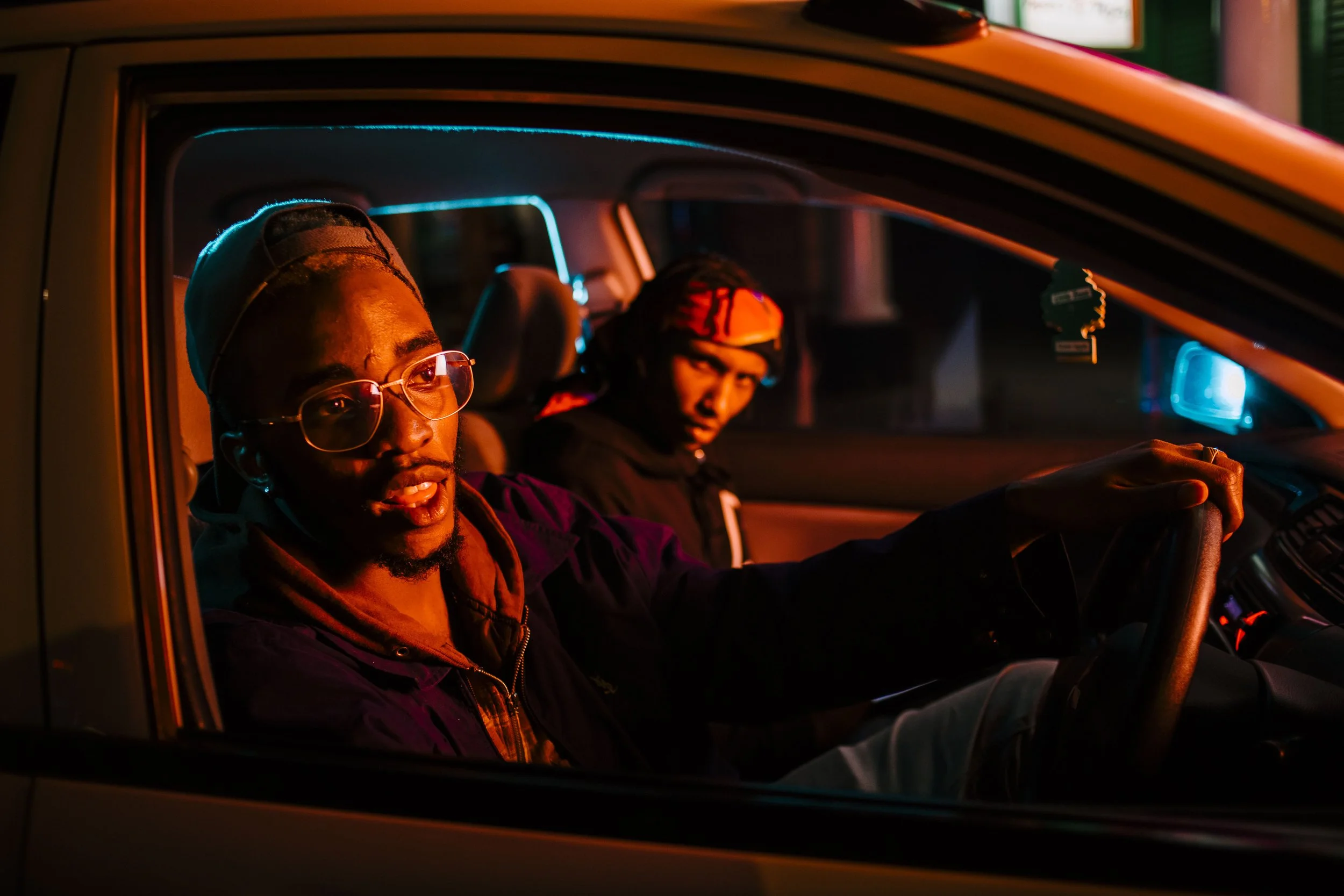 Two young men sitting in a car during nighttime, with one driving and the other in the passenger seat. The scene is illuminated by multicolored street lights, creating a vibrant, urban atmosphere.