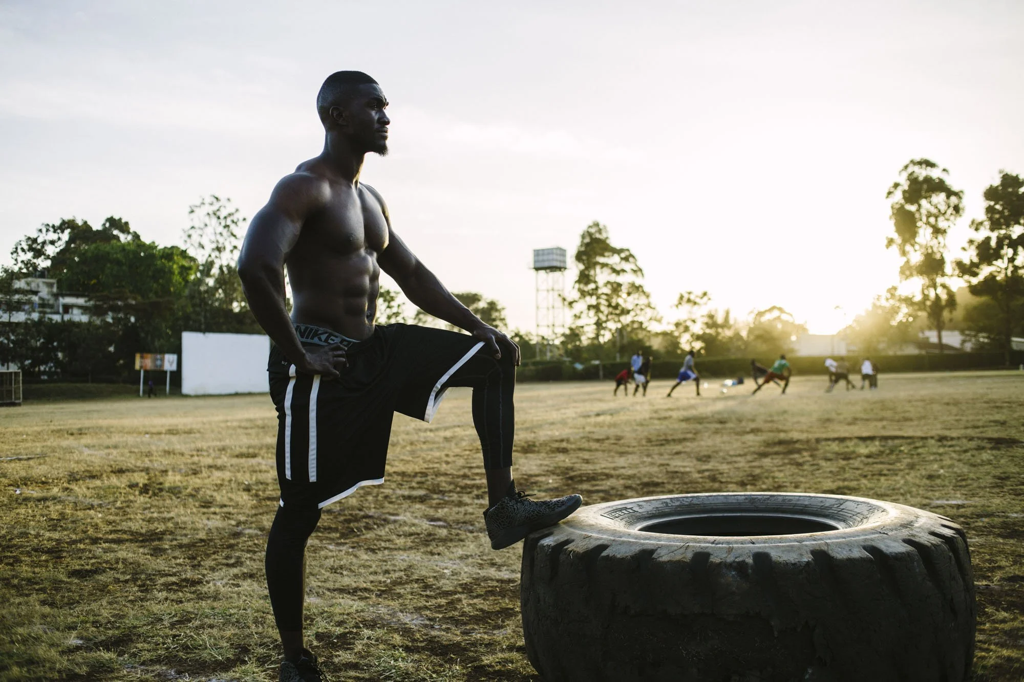 A shirtless man exercising outdoors at sunset, standing with one leg on a large tire and hands on his hips, with a group of people and trees in the background.