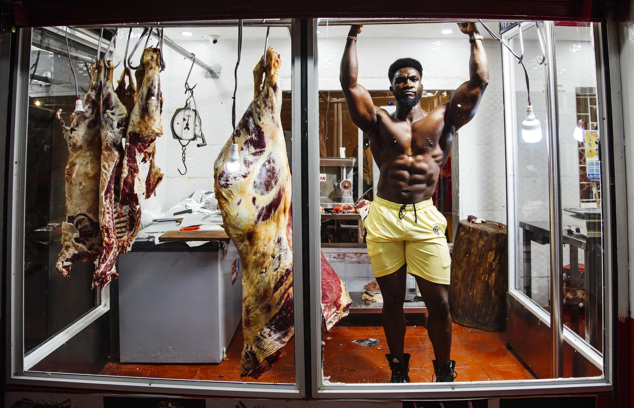 A muscular man lifting a barbell behind a glass window, with hanging meat carcasses in a butcher shop.