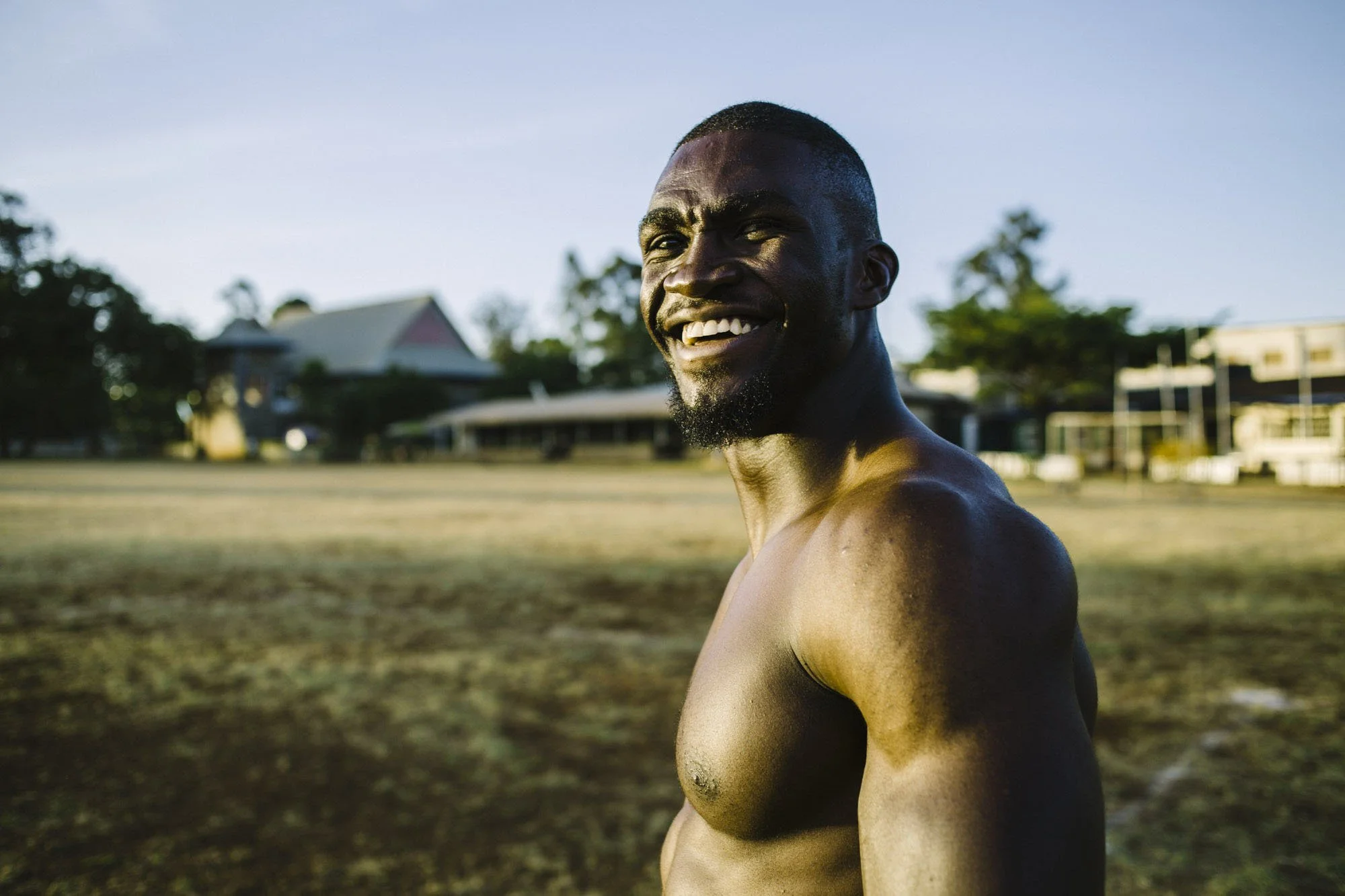 A shirtless smiling man with dark skin and a beard outdoors on a sunny day.