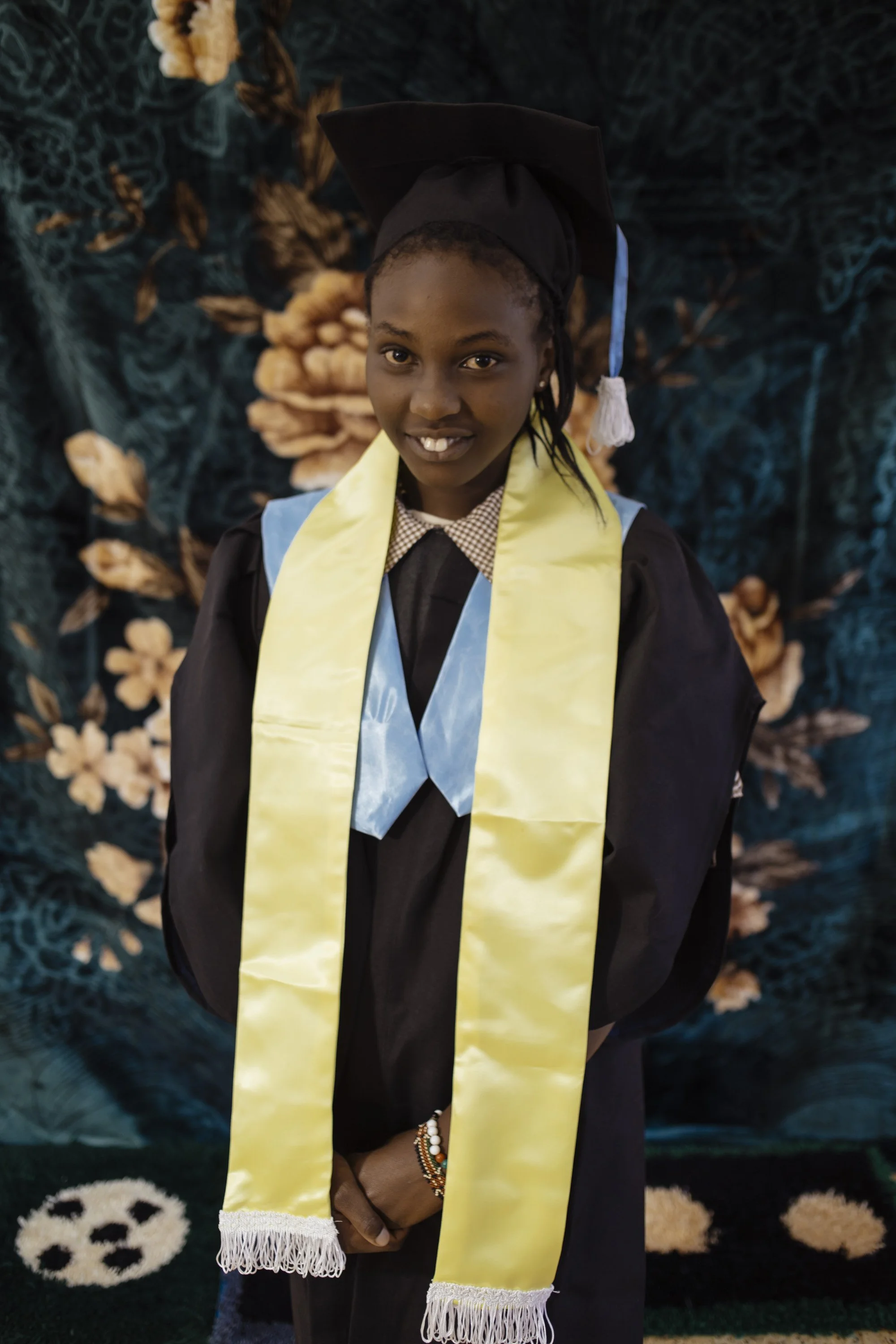 Young girl in graduation cap and gown, standing in front of a floral background, smiling with hands clasped.