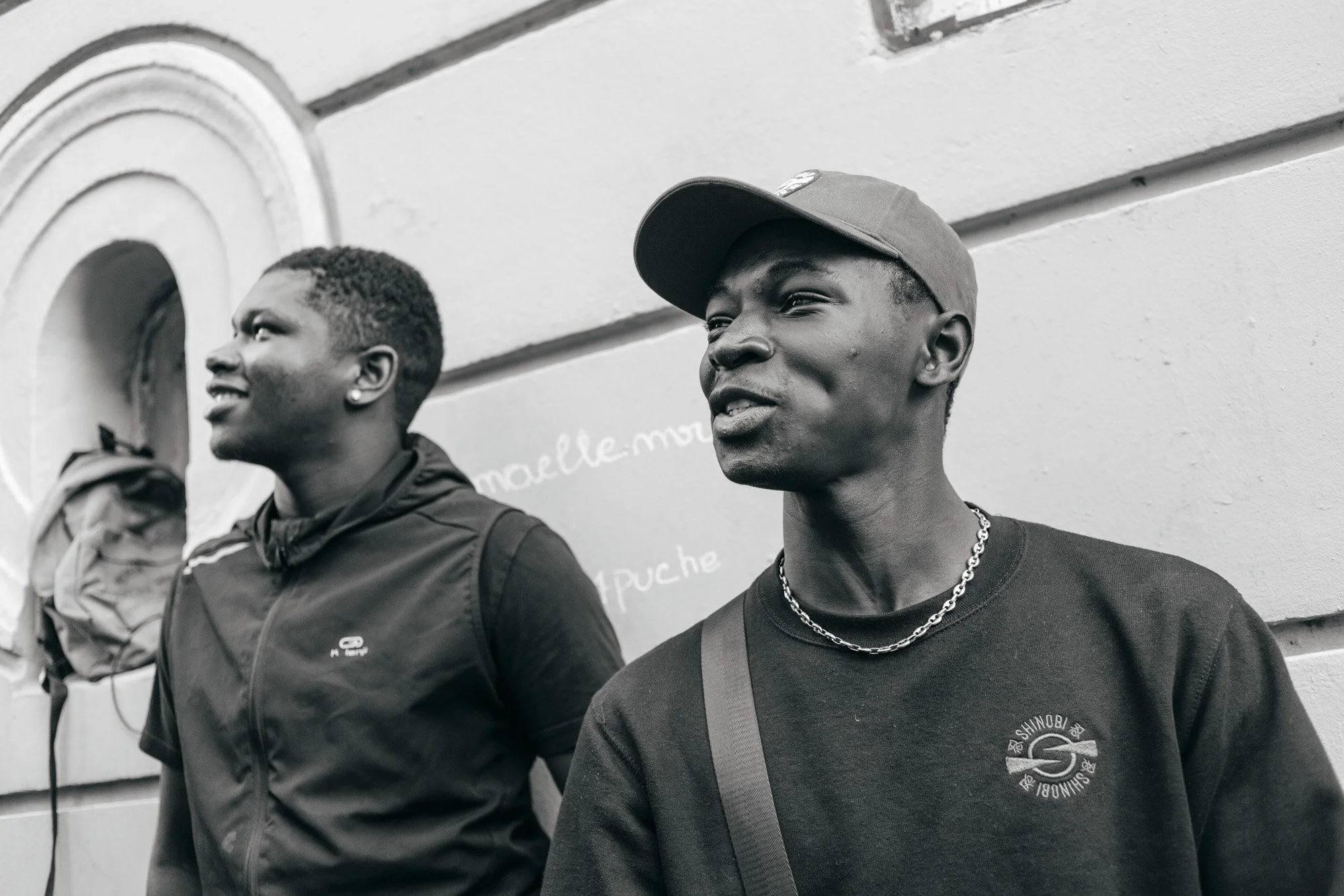 Two young men standing outdoors against a wall; one in a baseball cap and chain necklace, the other with a backpack, both looking to the side.