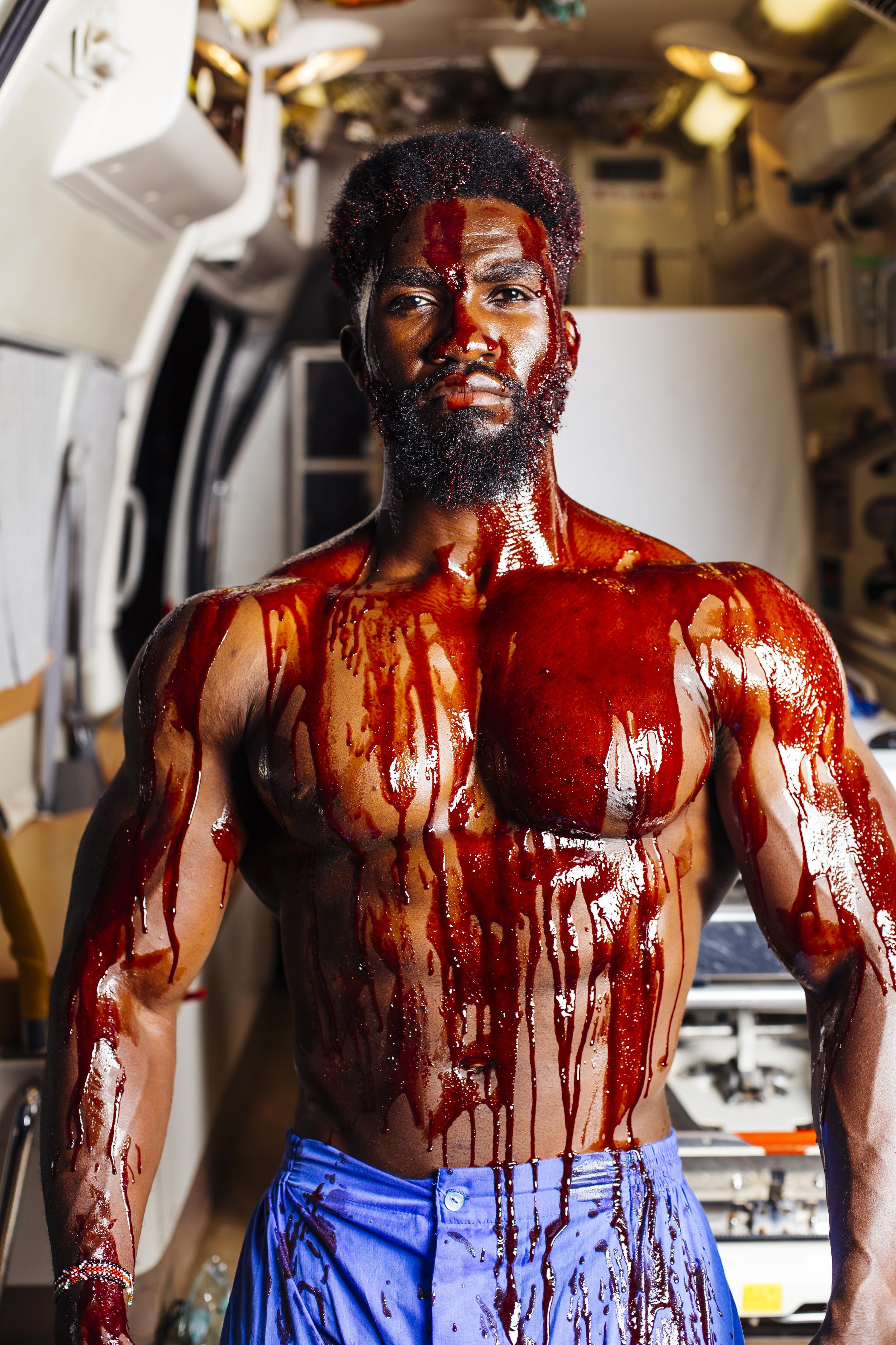 A man with blood-like substance on his face, chest, and arms in a confined space resembling a spacecraft or medical setting, wearing blue shorts.