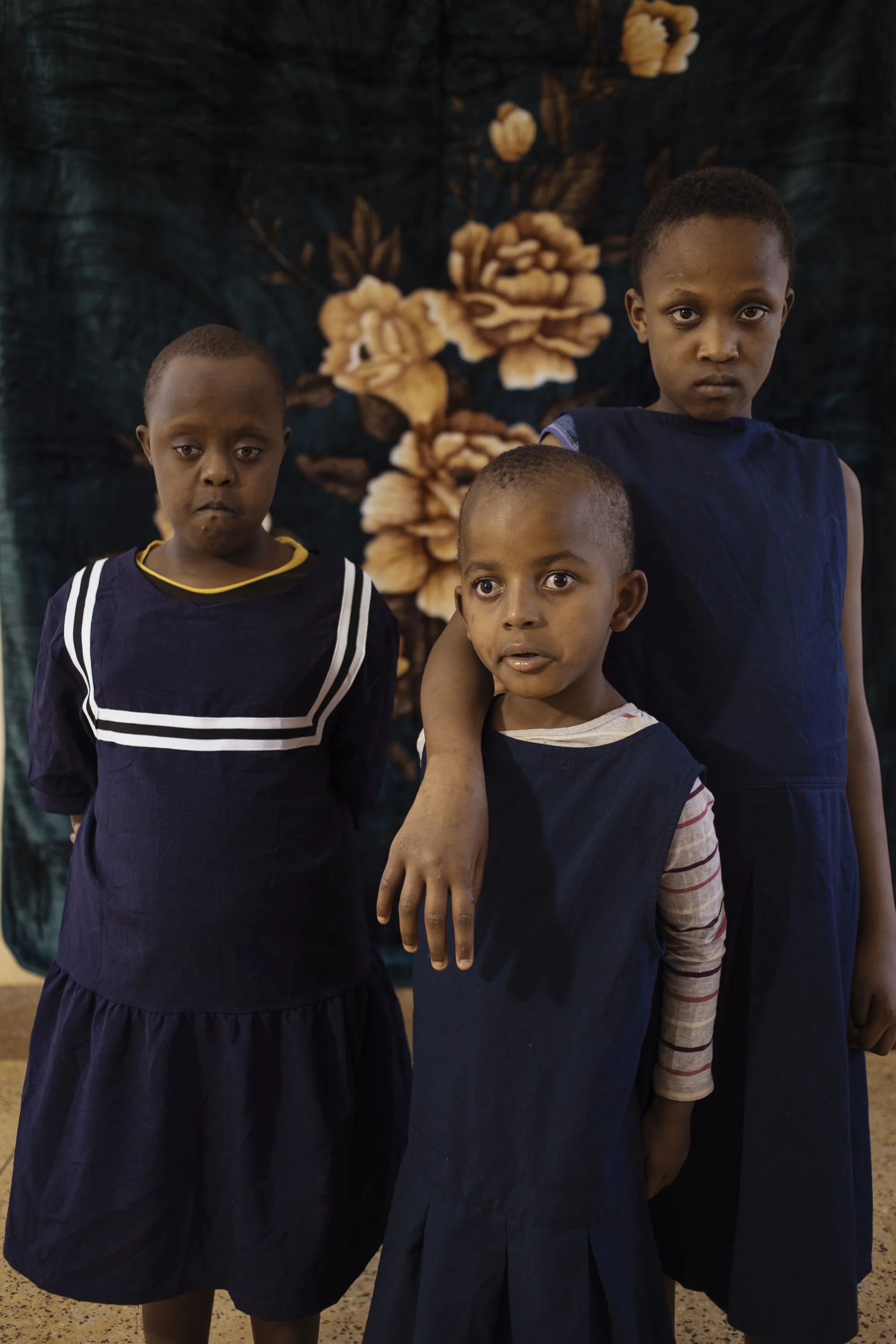 Three young children in navy blue dresses and shirts standing in front of a floral backdrop. One boy has his arm around a girl with short hair and expressive eyes, another girl stands beside them with a serious expression.