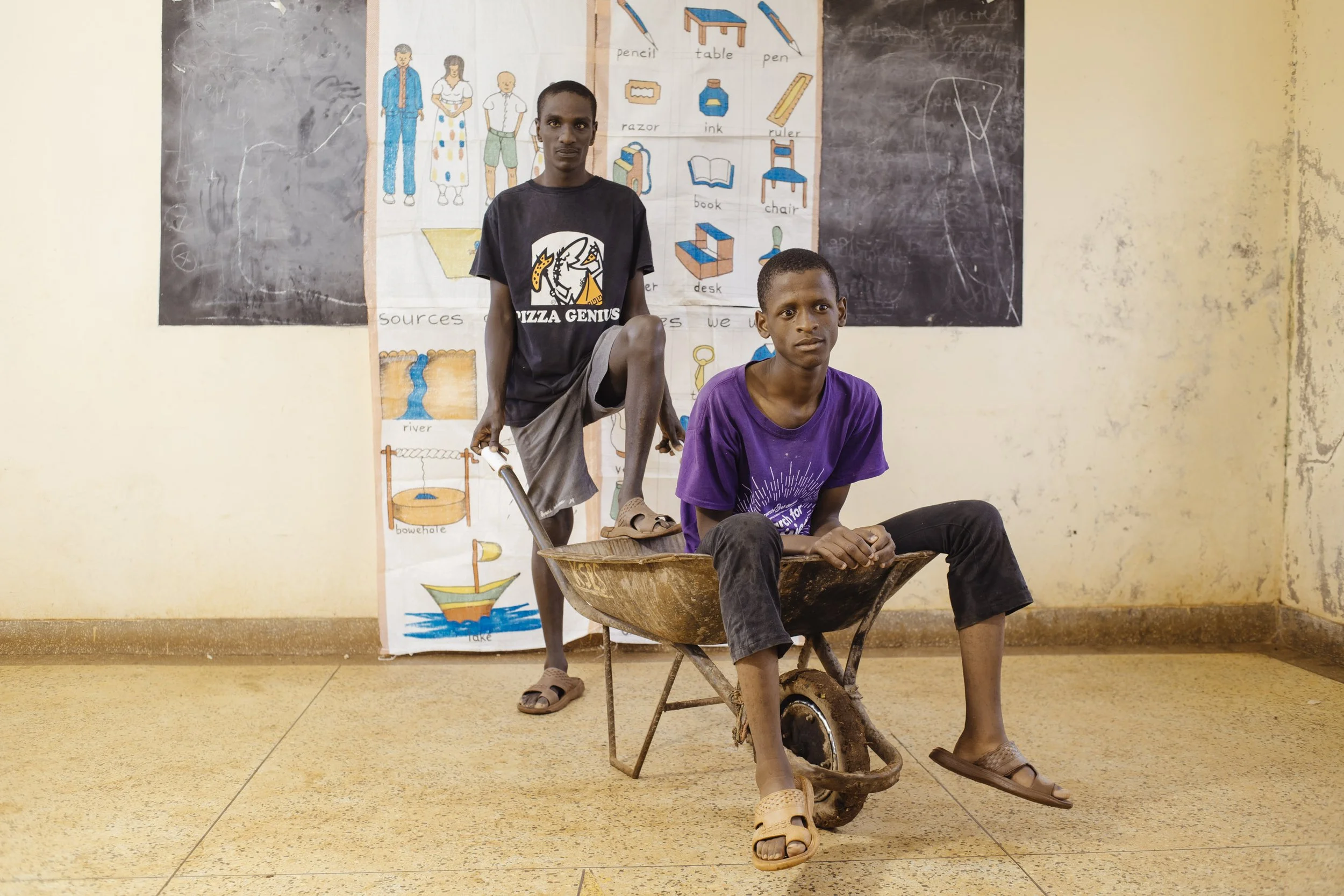 Two young boys in a classroom: one sitting in a wheelbarrow, the other standing behind it with one foot on the wheelbarrow. They are in front of an educational poster and blackboards.