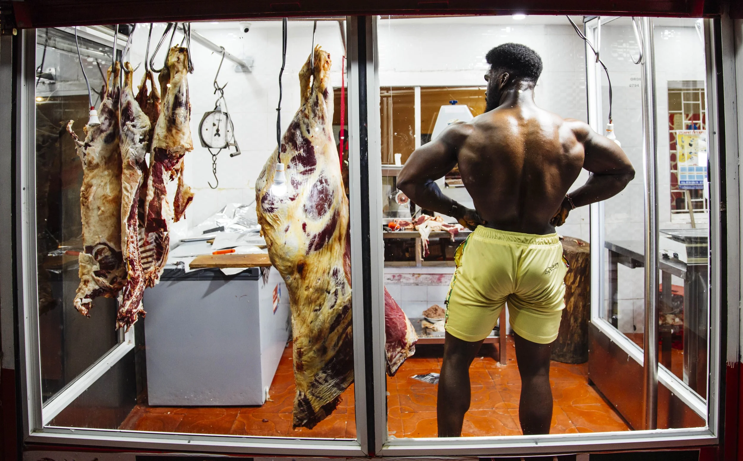 A muscular man stands shirtless in a butcher shop, looking at hanging meats inside a refrigerated display case.