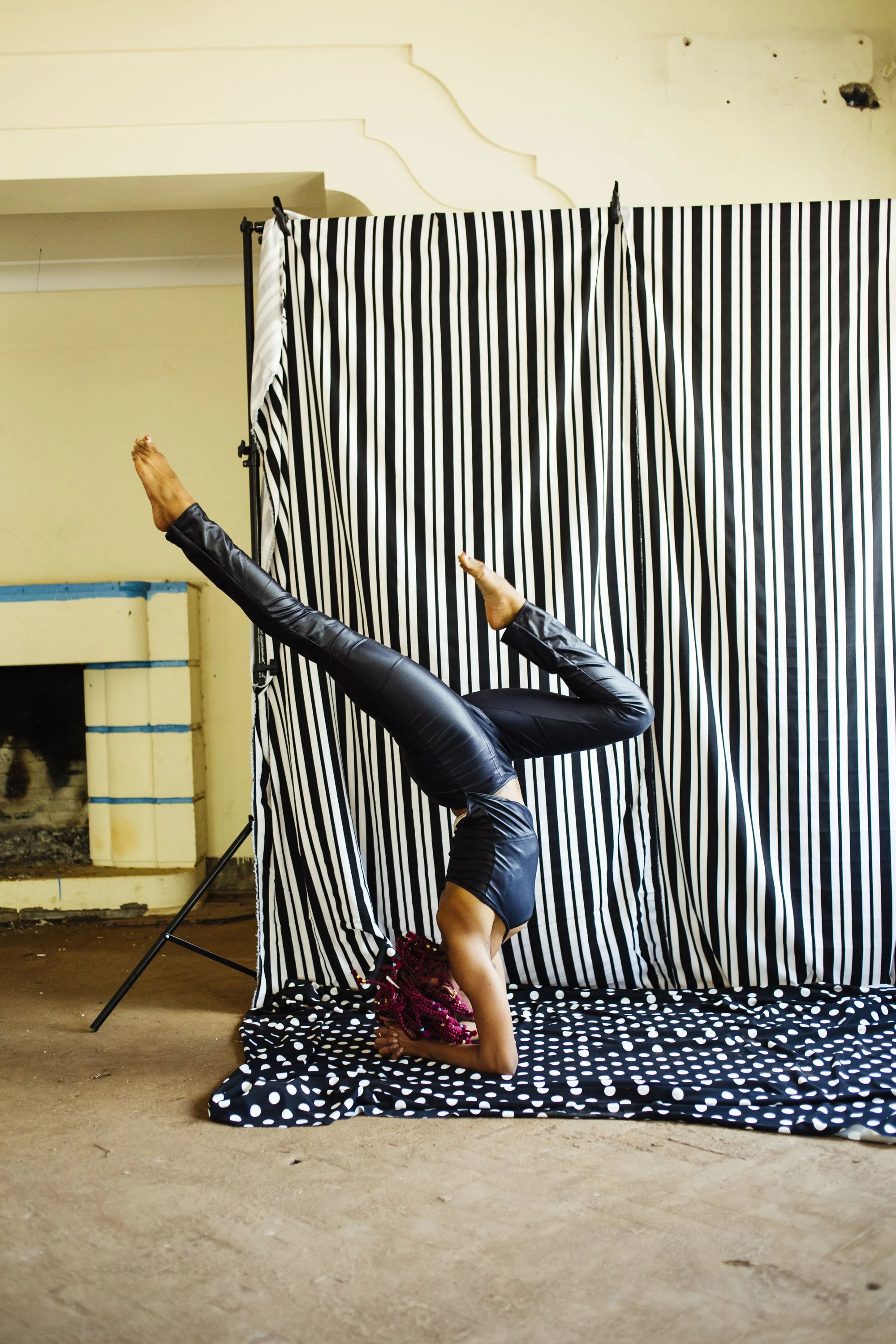 Person performing an upside-down yoga pose on a polka dot mat in front of black and white striped curtain.