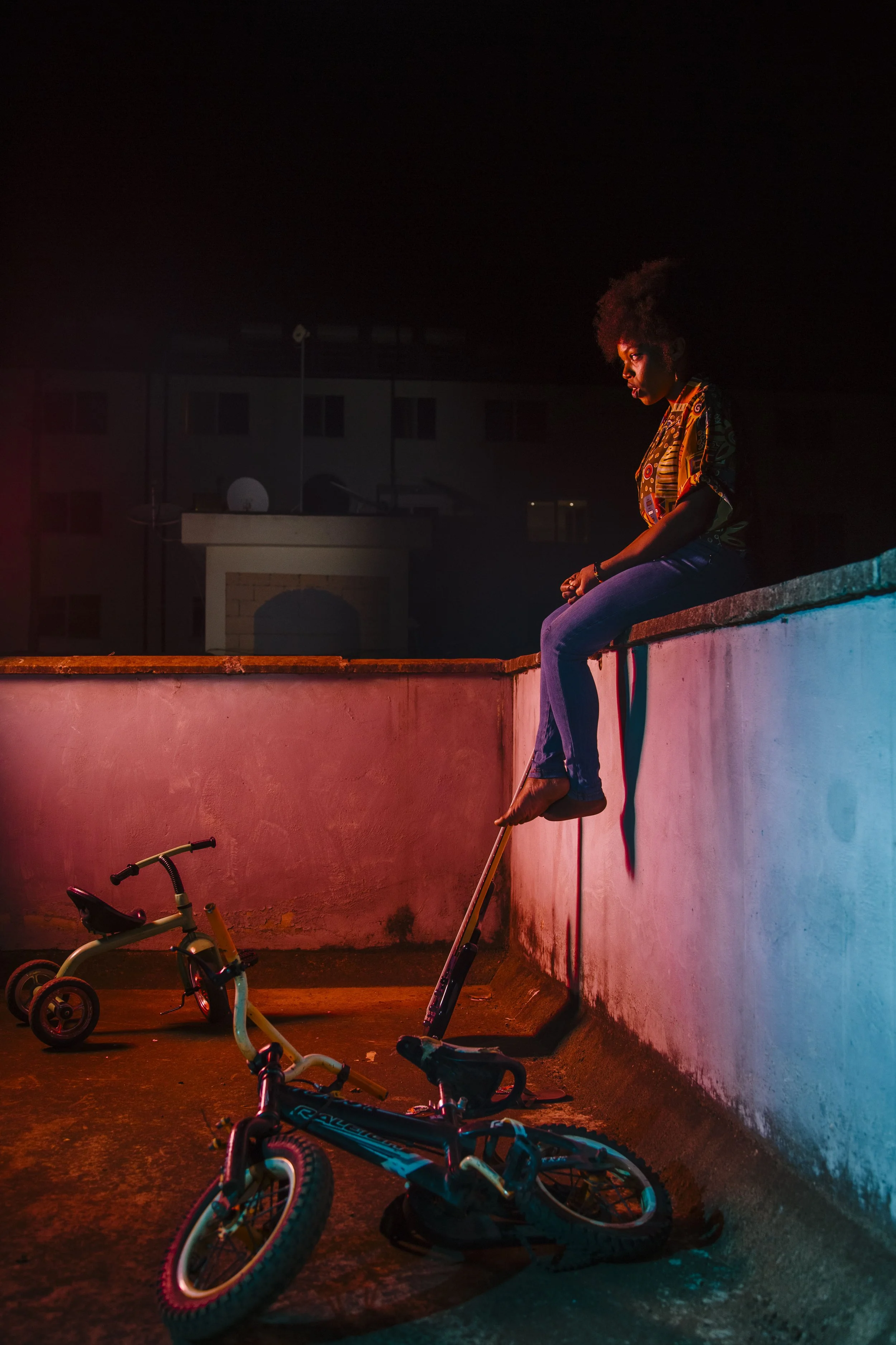 A young woman with curly hair sitting on the edge of a rooftop wall at night, illuminated by colorful neon lights, with a fallen bicycle and a children's tricycle on the floor.