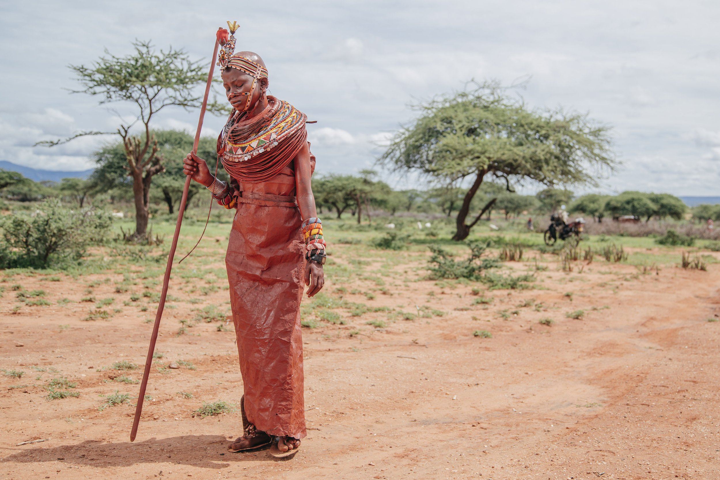 A Maasai woman dressed in traditional attire standing in a desert landscape with sparse trees and a cloudy sky, holding a staff.