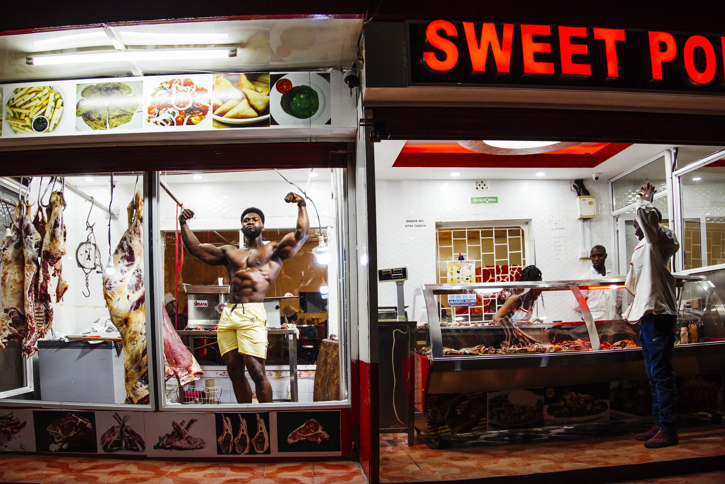 A butcher shop with hanging meat, a shirtless man flexing muscles inside the butcher shop, and a customer talking to the staff outside at the meat counter.