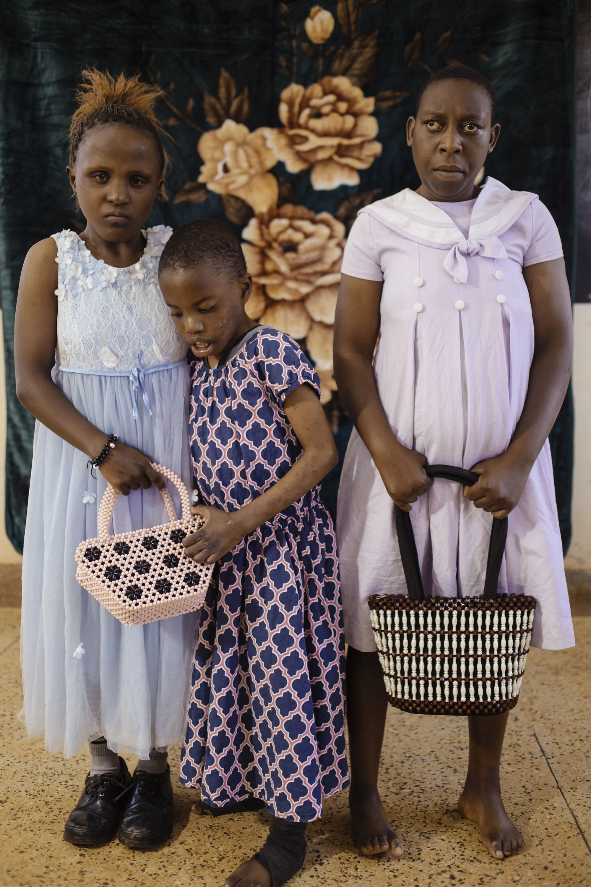 Three women standing in front of a floral backdrop, holding handbags, with serious expressions.
