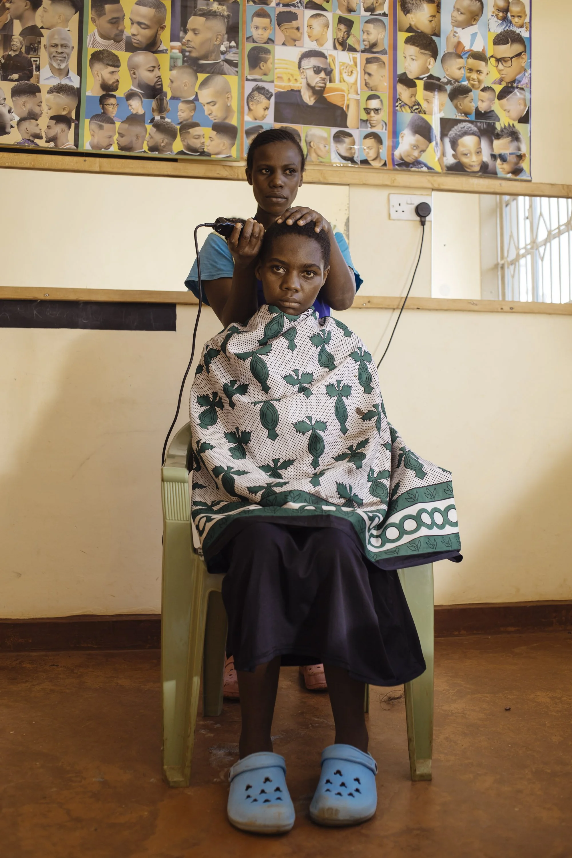 A young boy seated in a green plastic chair gets a haircut by a woman standing behind him. The boy is wearing a patterned cape and blue Crocs, and the woman is cutting his hair with an electric clipper. The background features a collage of various ha