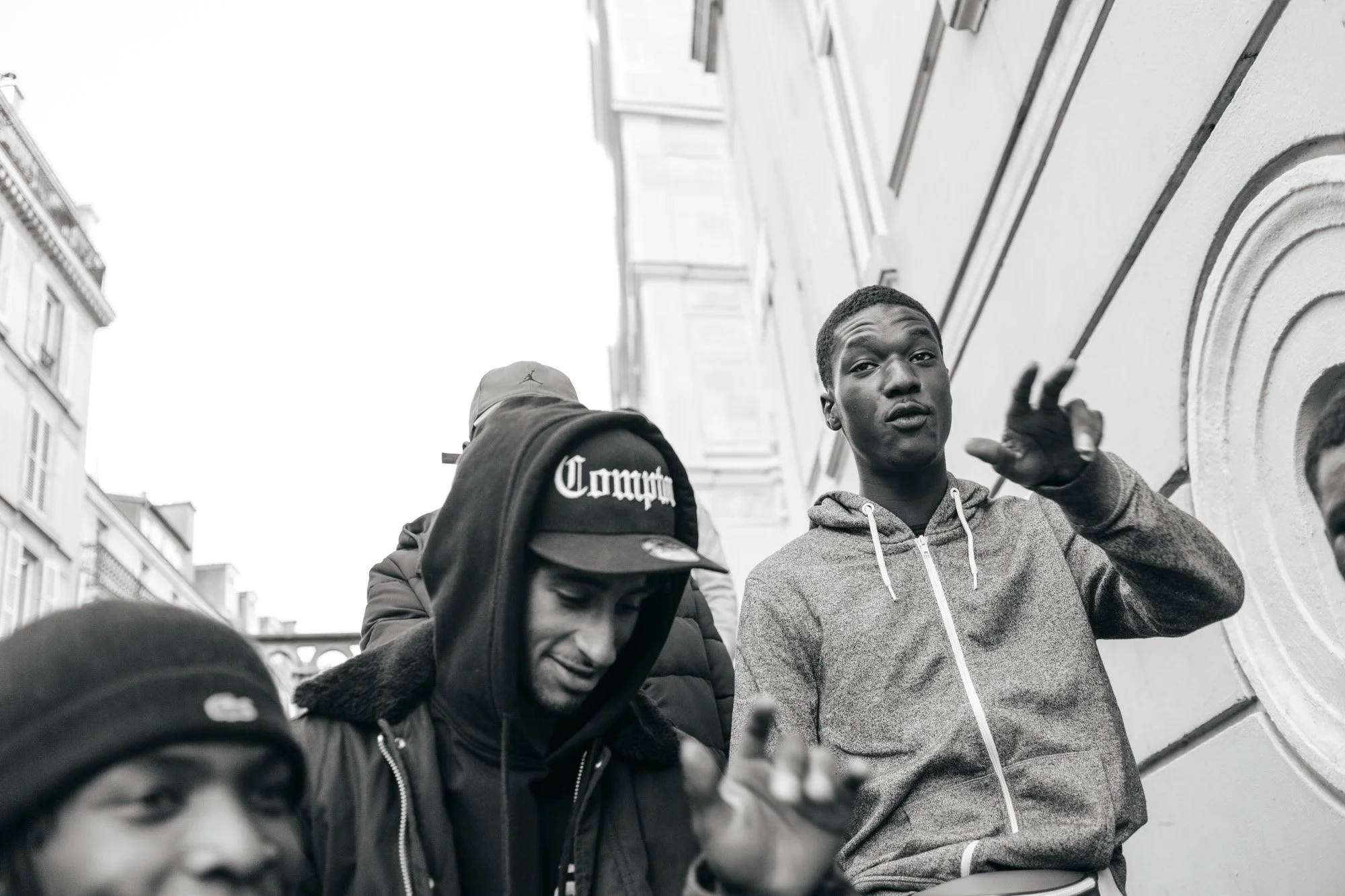 Group of young men, one speaking with hand raised, outside urban building in black and white.