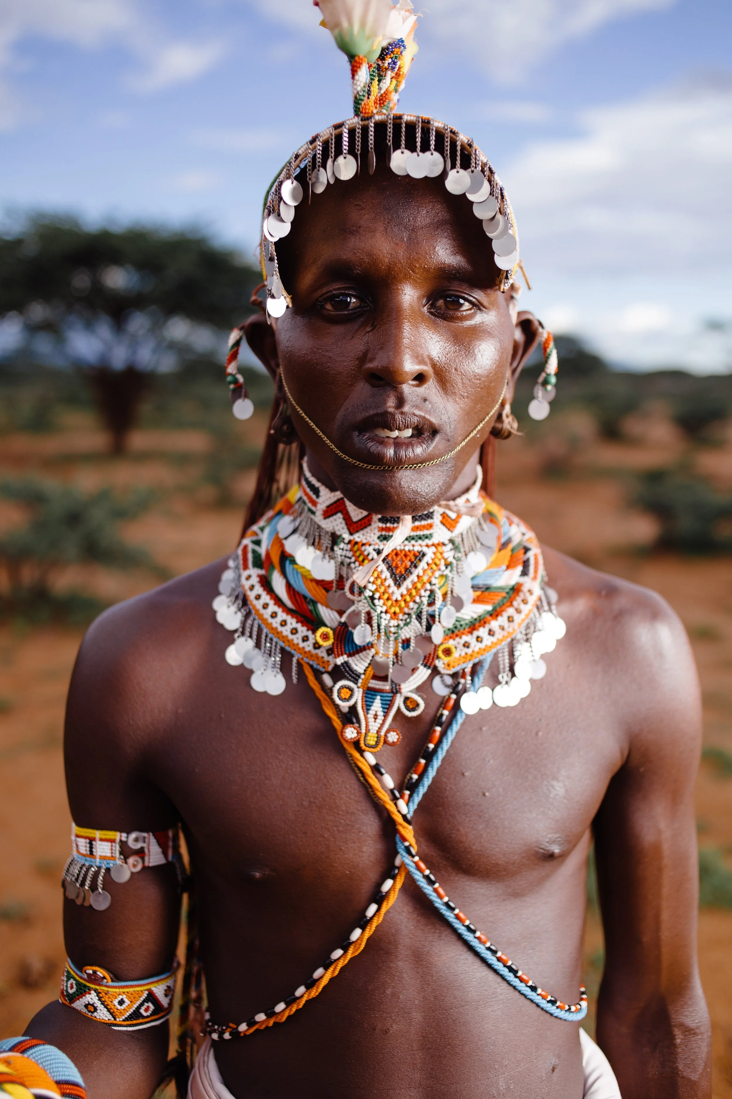 A person from an African tribe wearing traditional colorful bead jewelry and accessories, with a natural outdoor background of trees and sky.