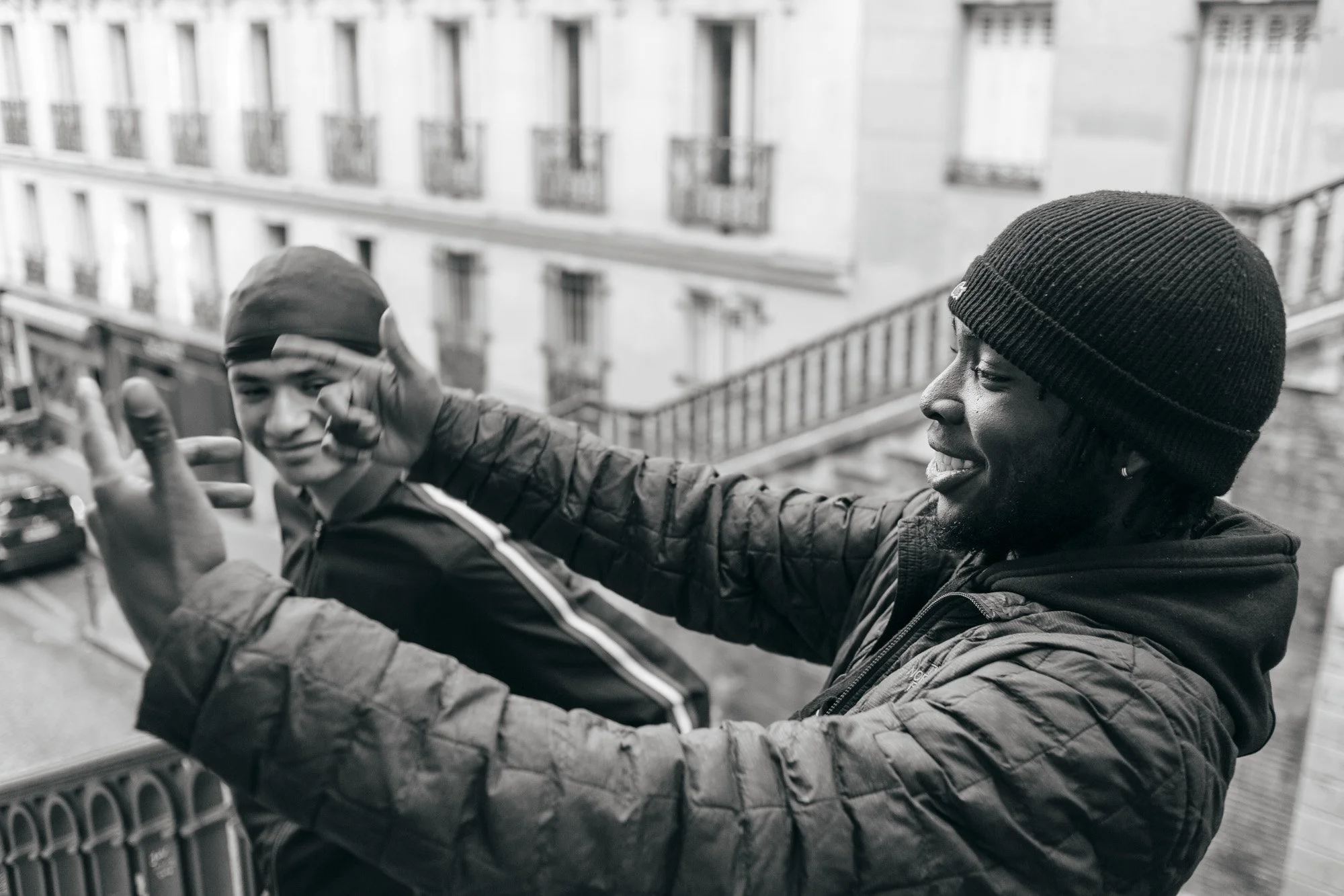 Two young men with dark skin enjoying themselves on a city balcony, with buildings and staircases in the background. One man wears a beanie, the other a cap. They are smiling and making hand gestures.