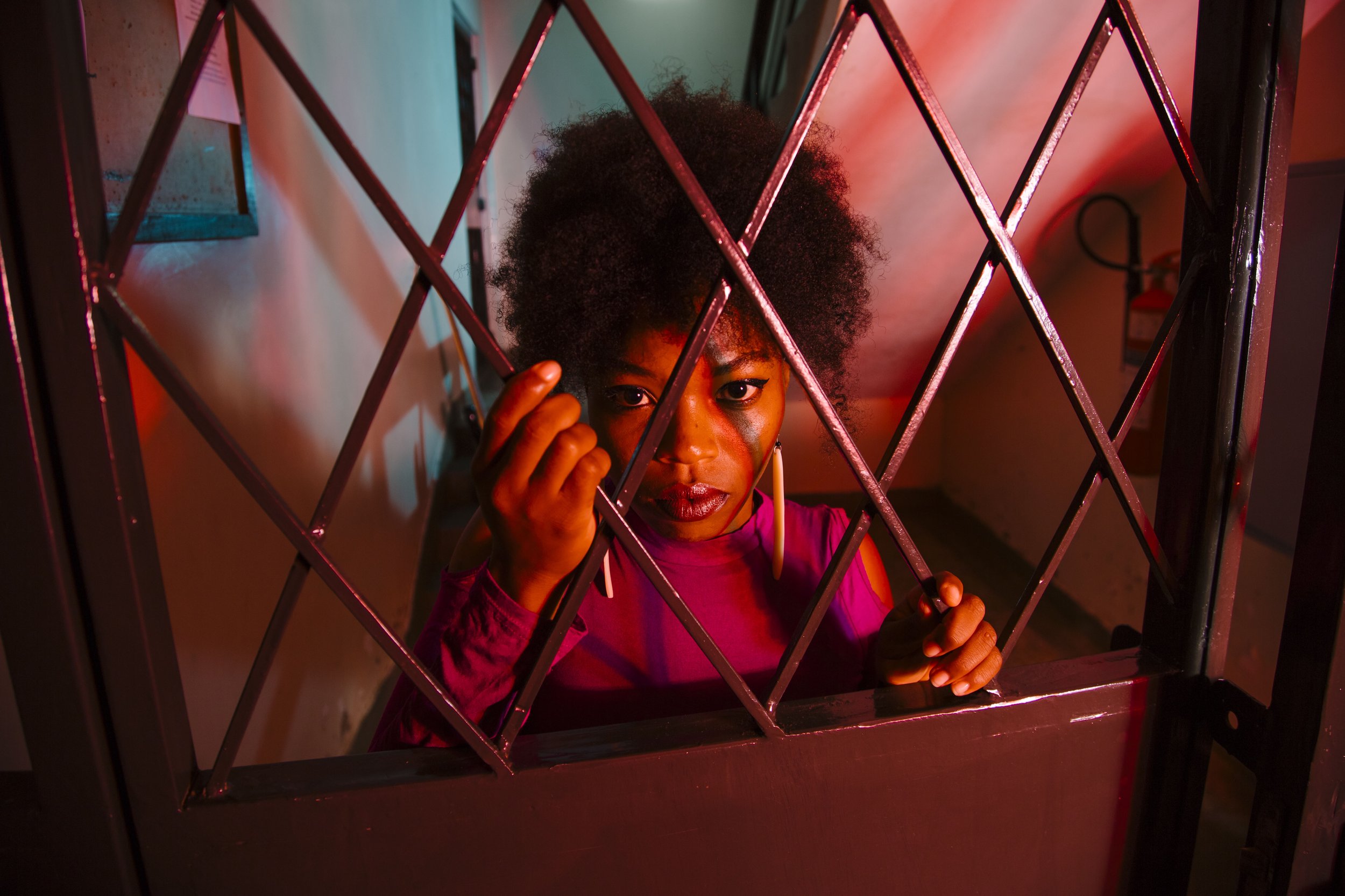 A woman with curly hair looking through a metal gate, illuminated by red and blue lighting.
