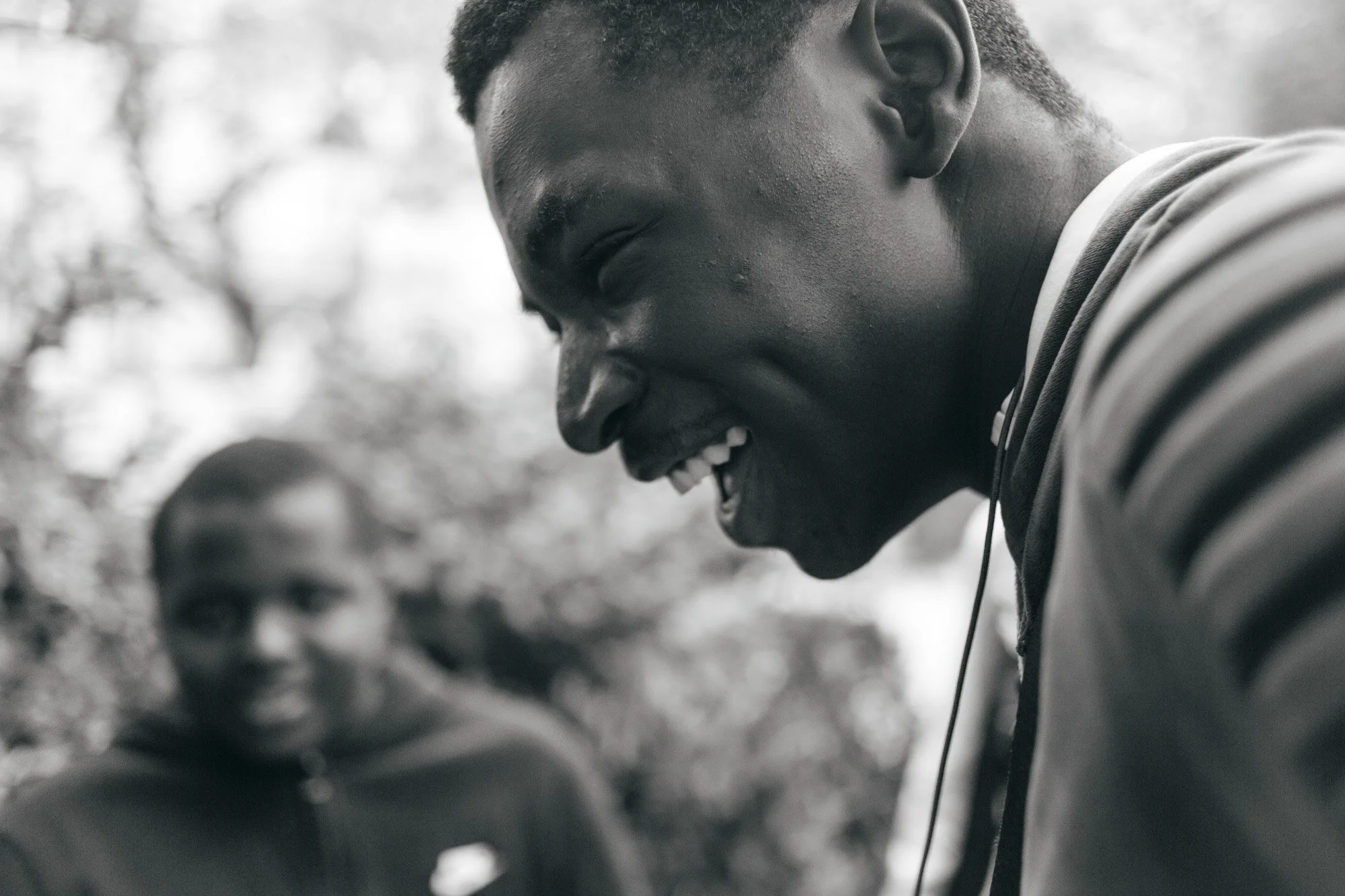 Smiling African American male outdoors, wearing a striped shirt, with blurred person in background.
