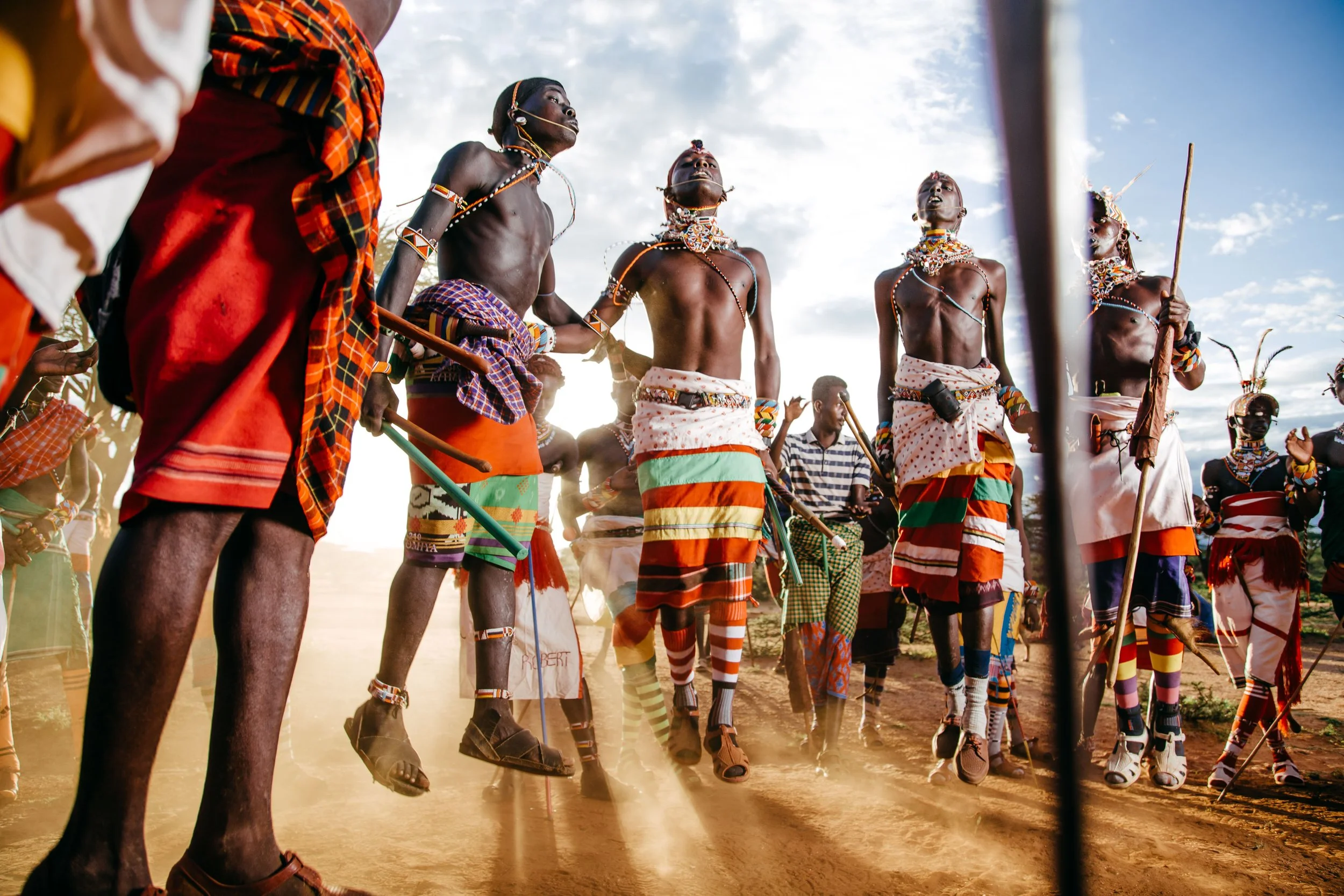 Group of indigenous men participating in a traditional dance or ceremony outdoors, dressed in colorful clothing and accessories, with some holding sticks.