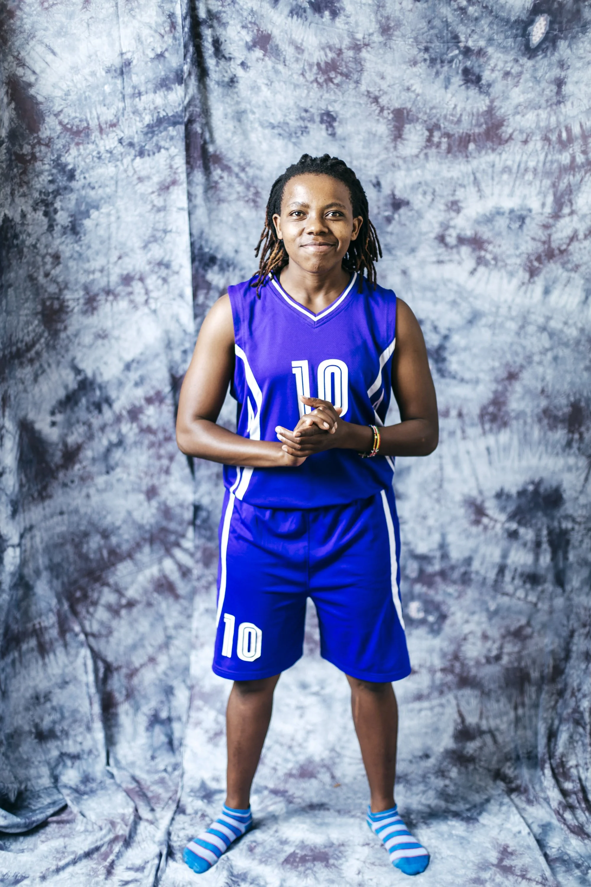 Young girl in a blue basketball uniform with the number 10, standing with hands clasped, standing on a tie-dye styled backdrop, wearing striped socks.