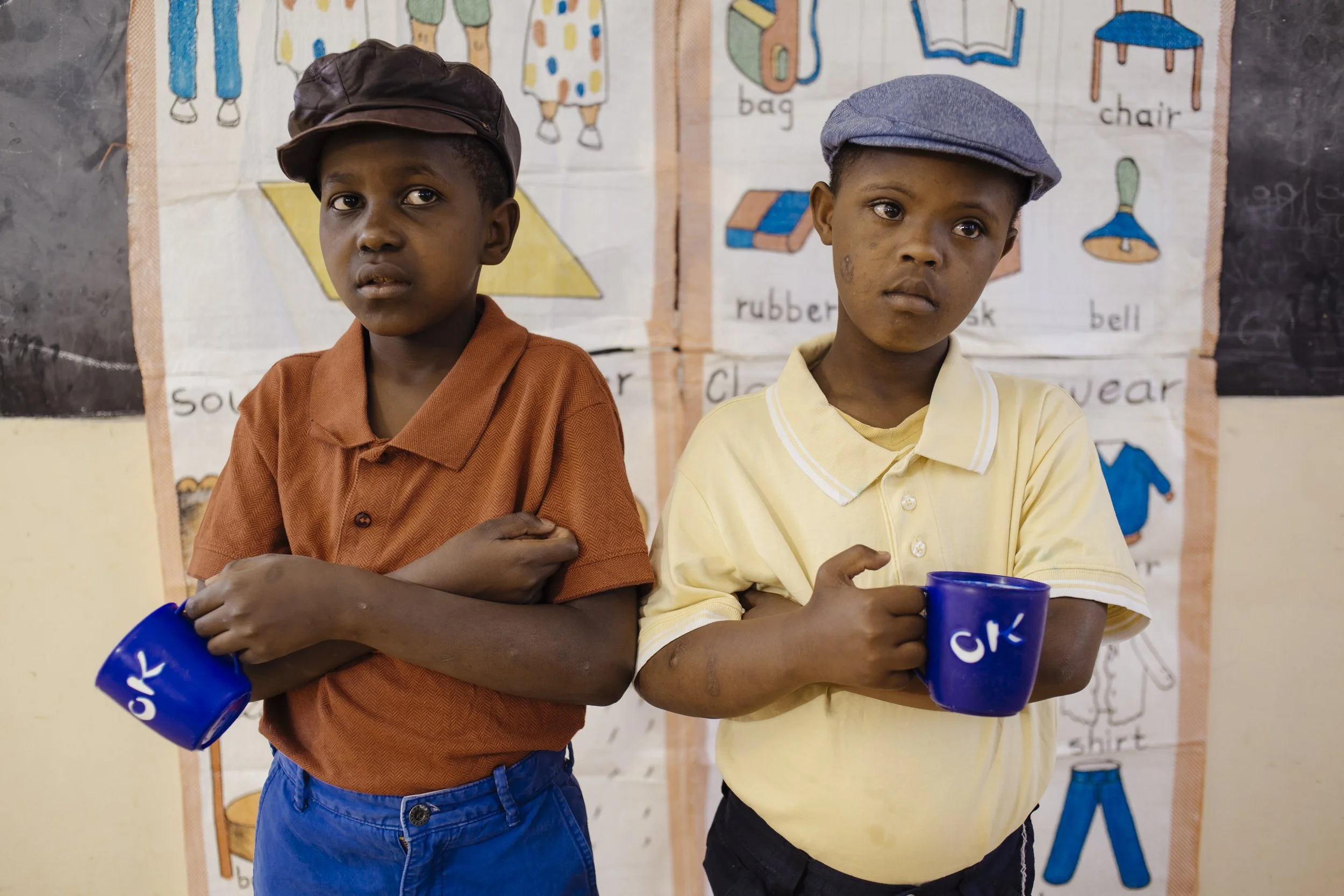 Two young boys standing in a classroom, holding blue mugs with the initials 'CK' on them. They appear to be students, with a colorful educational poster of classroom objects visible in the background.