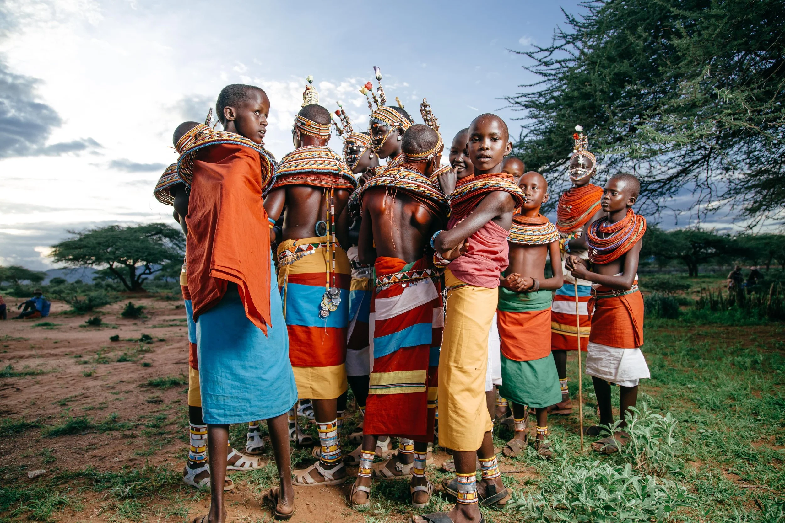 Group of Maasai children wearing traditional colorful jewelry and clothing, standing on a grassy landscape with trees under a cloudy sky.