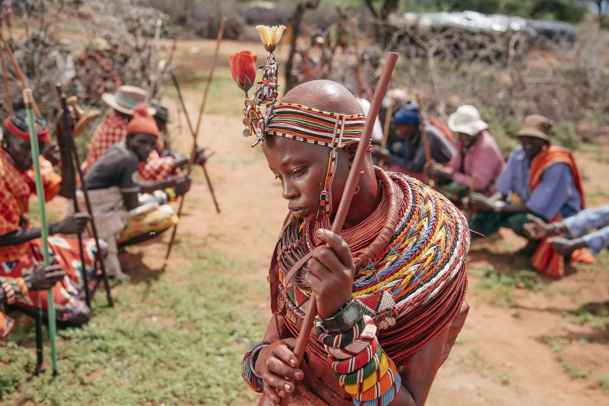 A woman from the Maasai tribe wearing traditional beadwork and jewelry, holding a wooden stick, with a group of Maasai people sitting on the ground in the background.