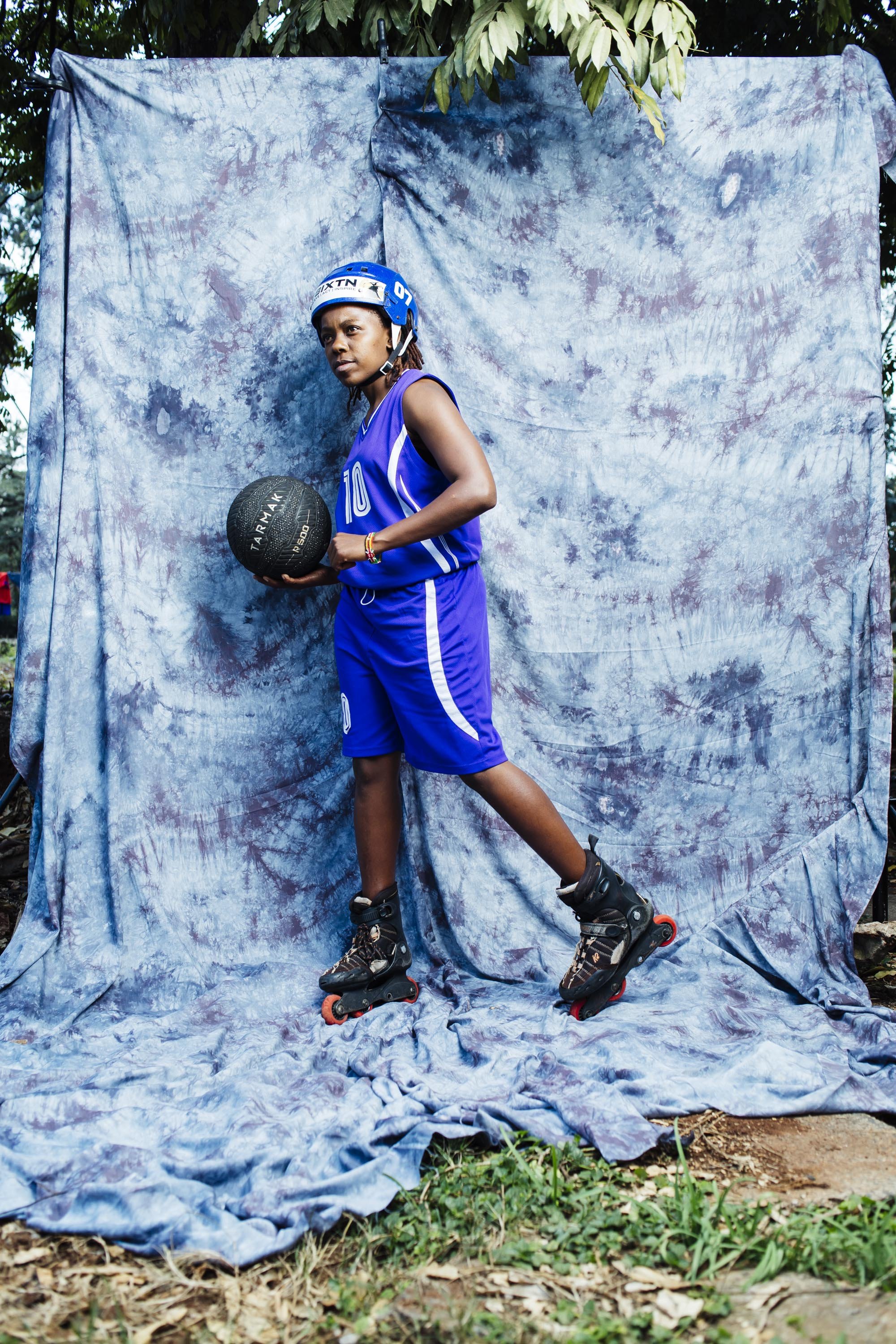 Young girl in blue basketball uniform and rollerblades holding a basketball, standing outdoors against a blue tie-dye backdrop.