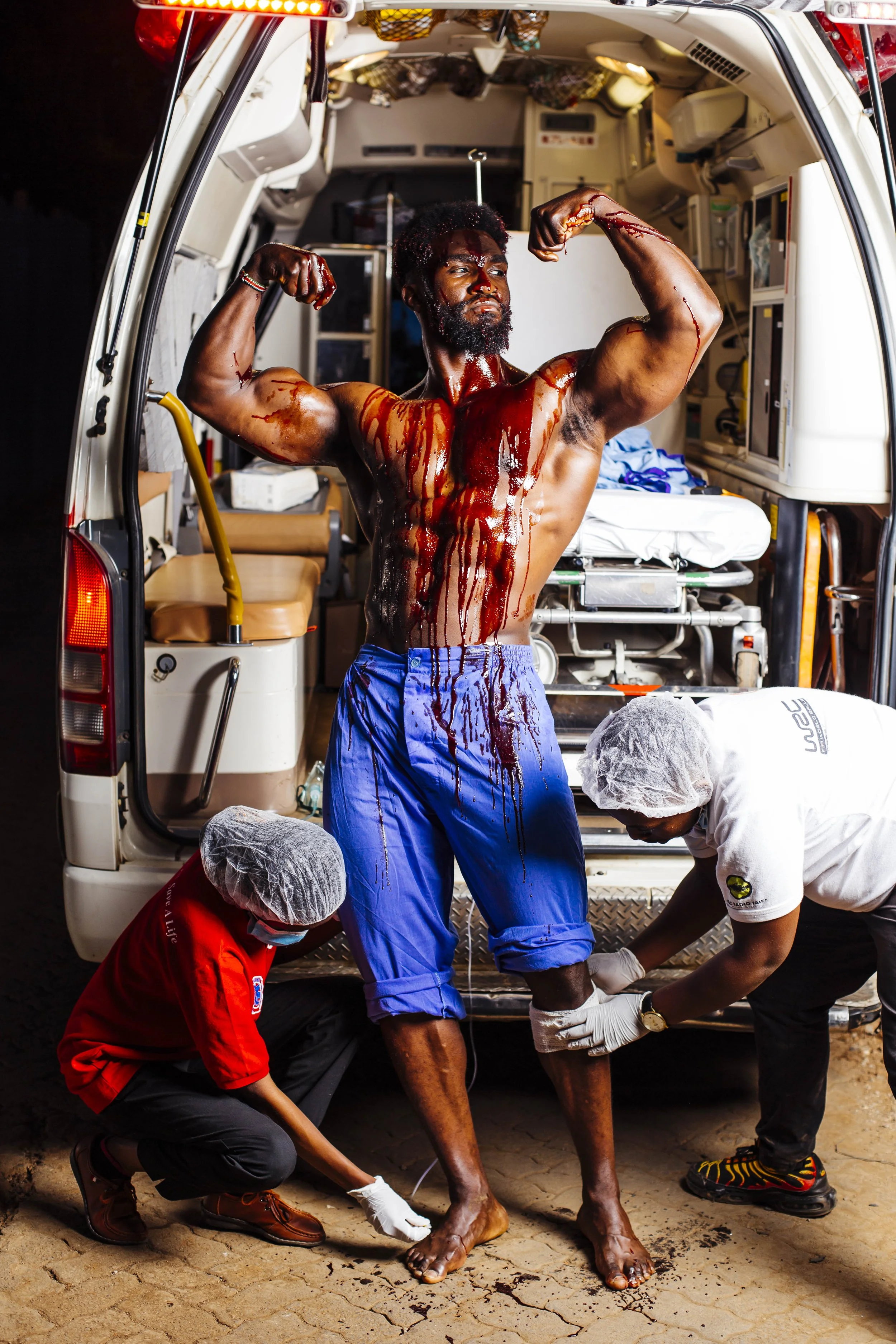 A man with blood on his chest and face is standing outside a medical vehicle, flexing his muscles. Two healthcare workers are attending to his legs, wearing gloves and hair covers.