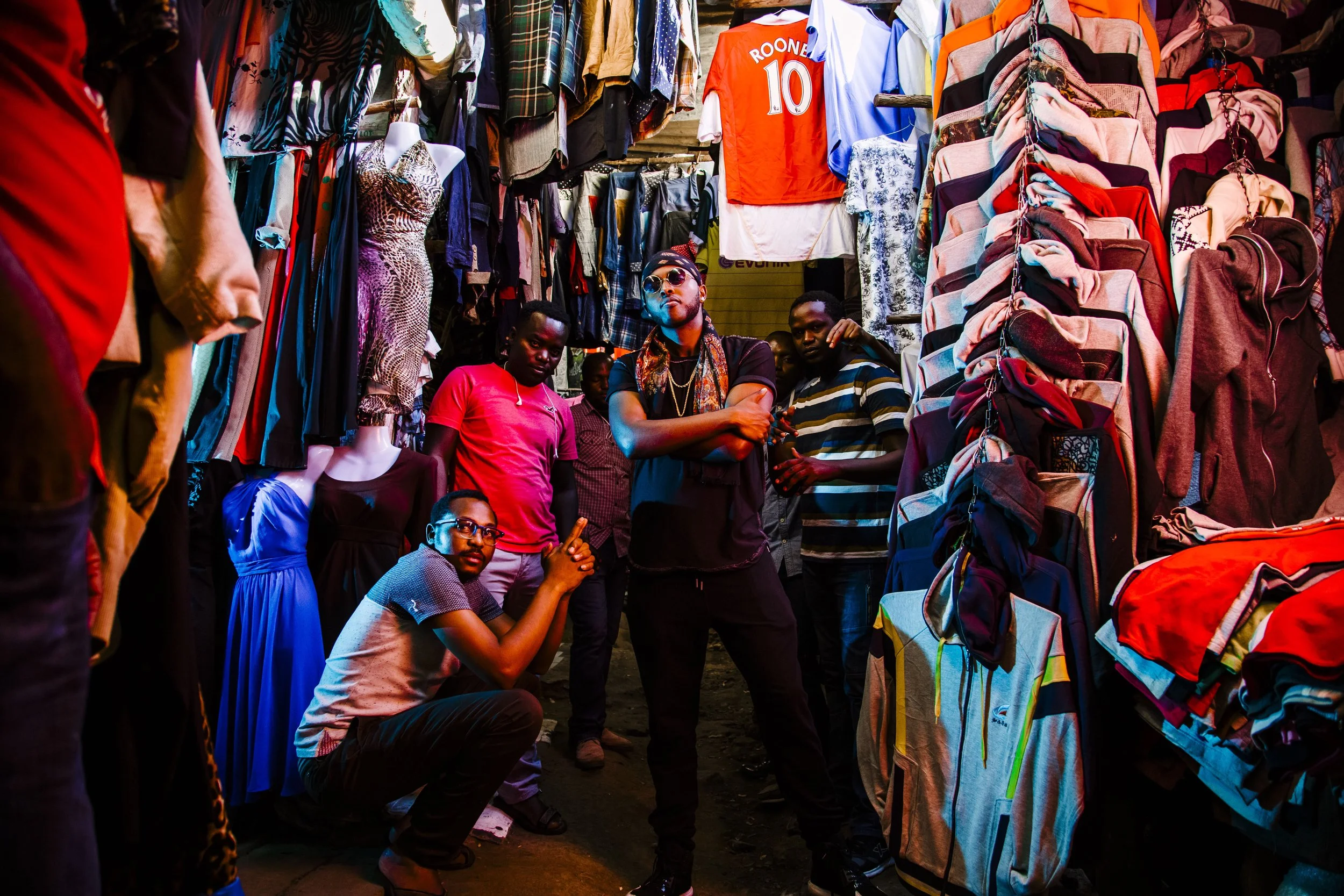 Group of young men in a busy market surrounded by hanging clothing and accessories, with colorful lighting.