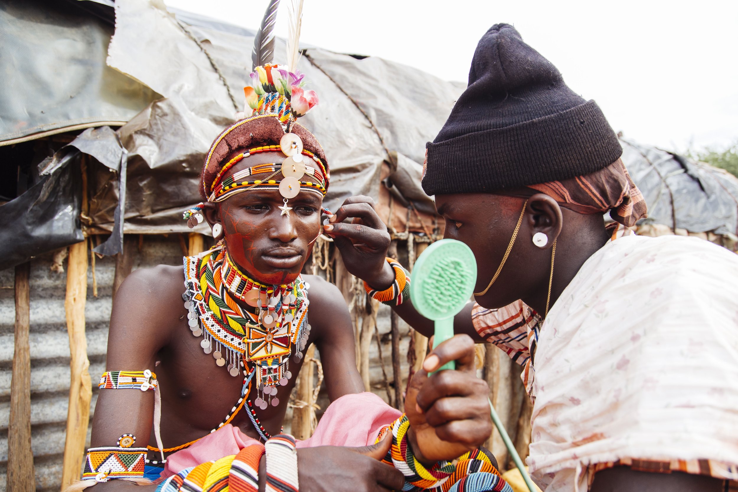 A young man dressed in traditional Maasai attire sitting still as a woman applies face paint with a small brush in front of a makeshift shelter.