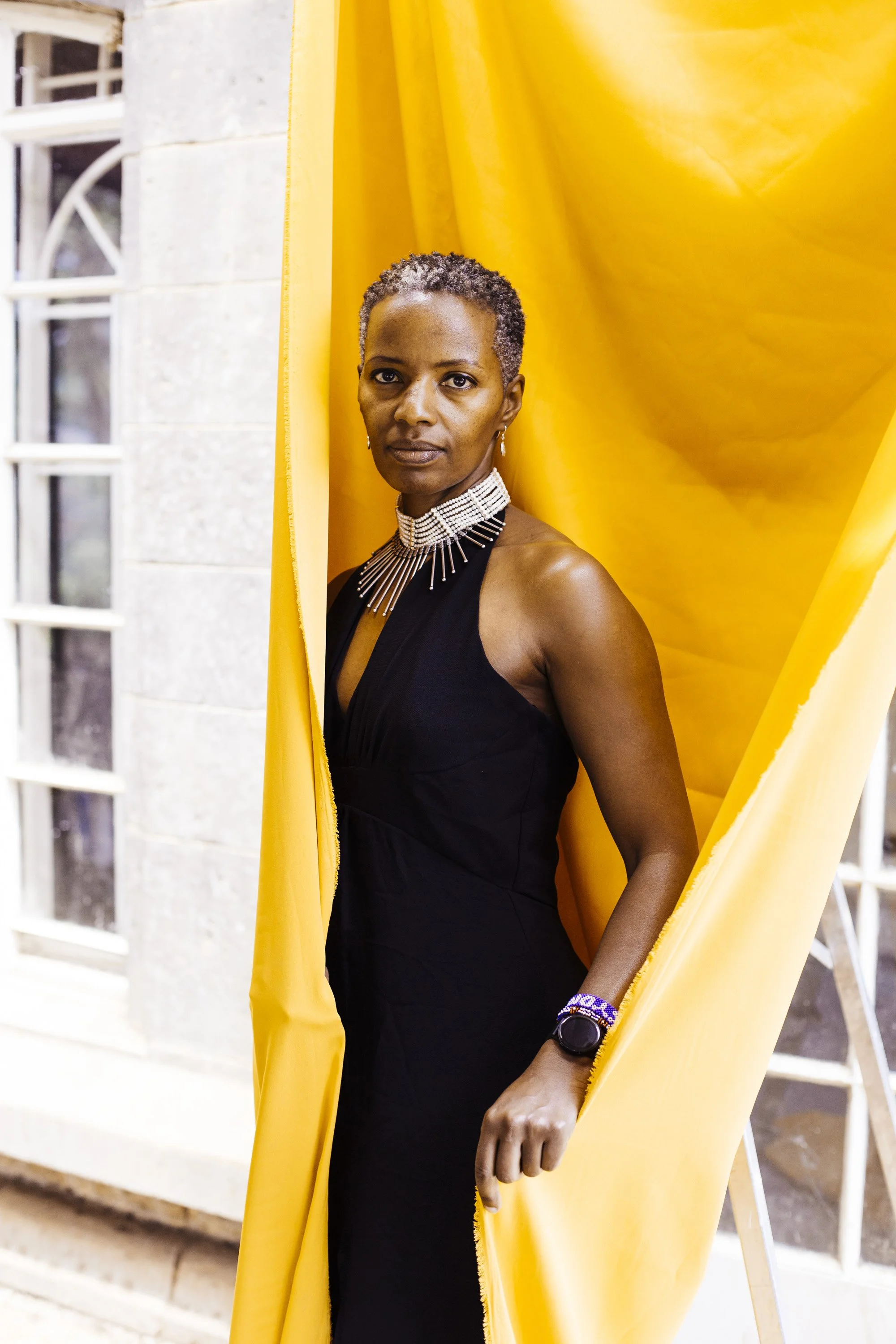 A woman with short gray hair standing behind a yellow curtain, wearing a black dress and a statement necklace, with a wall and window in the background.