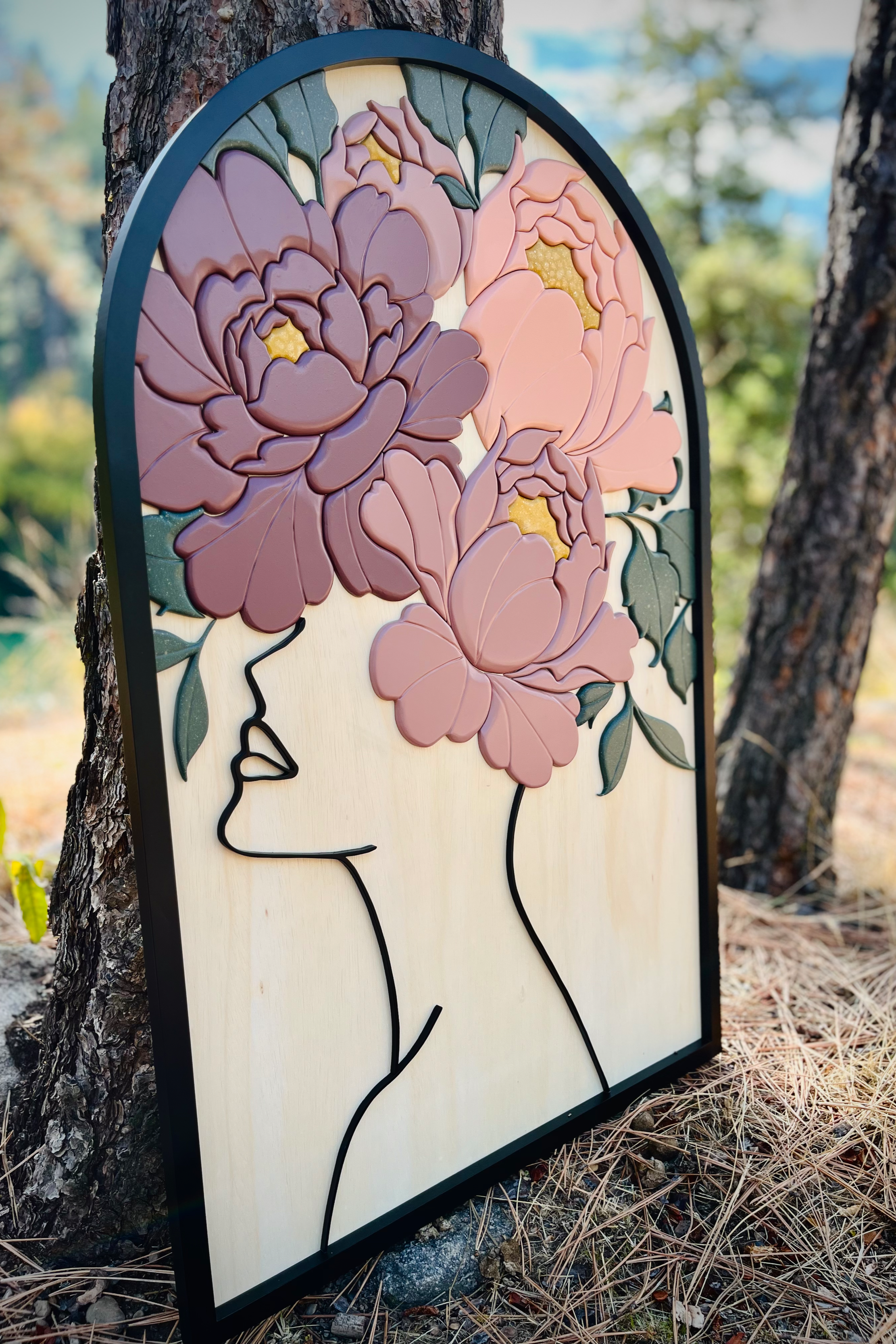 Artistic wood wall hanging of a woman's face in profile with her hair made of large pink and purple flowers and green leaves, set against a wooded outdoor background.