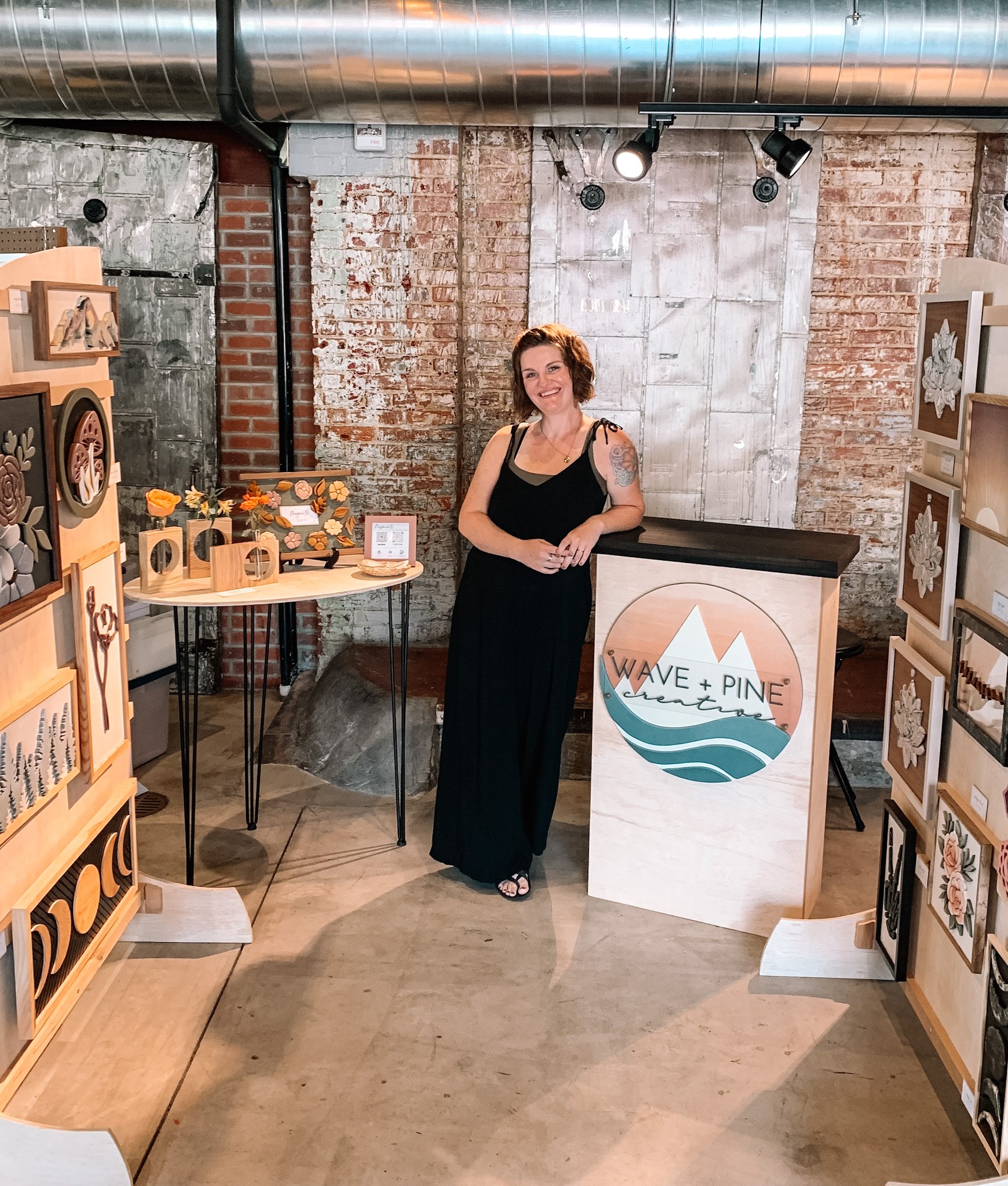 A woman standing behind a reception desk with a sign that says "Wave + Pine Creations". She is wearing a black dress and is smiling. There are art pieces and decorations on display to her left and right, with a rustic brick wall in the background.