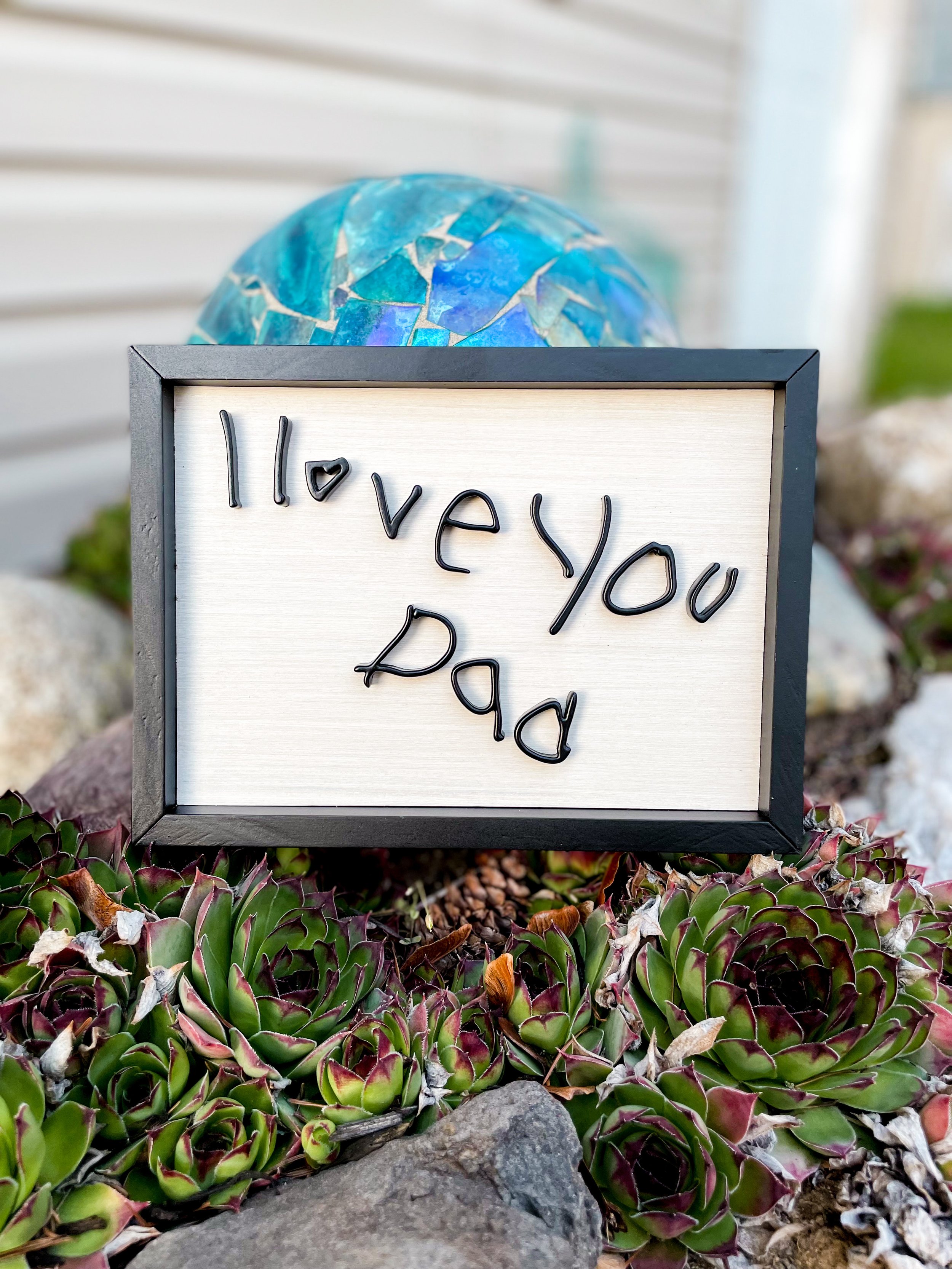 A sign that reads 'I love you Dad' with the words spelled out in black wire, placed among succulents and rocks, with a mosaic sphere in the background.