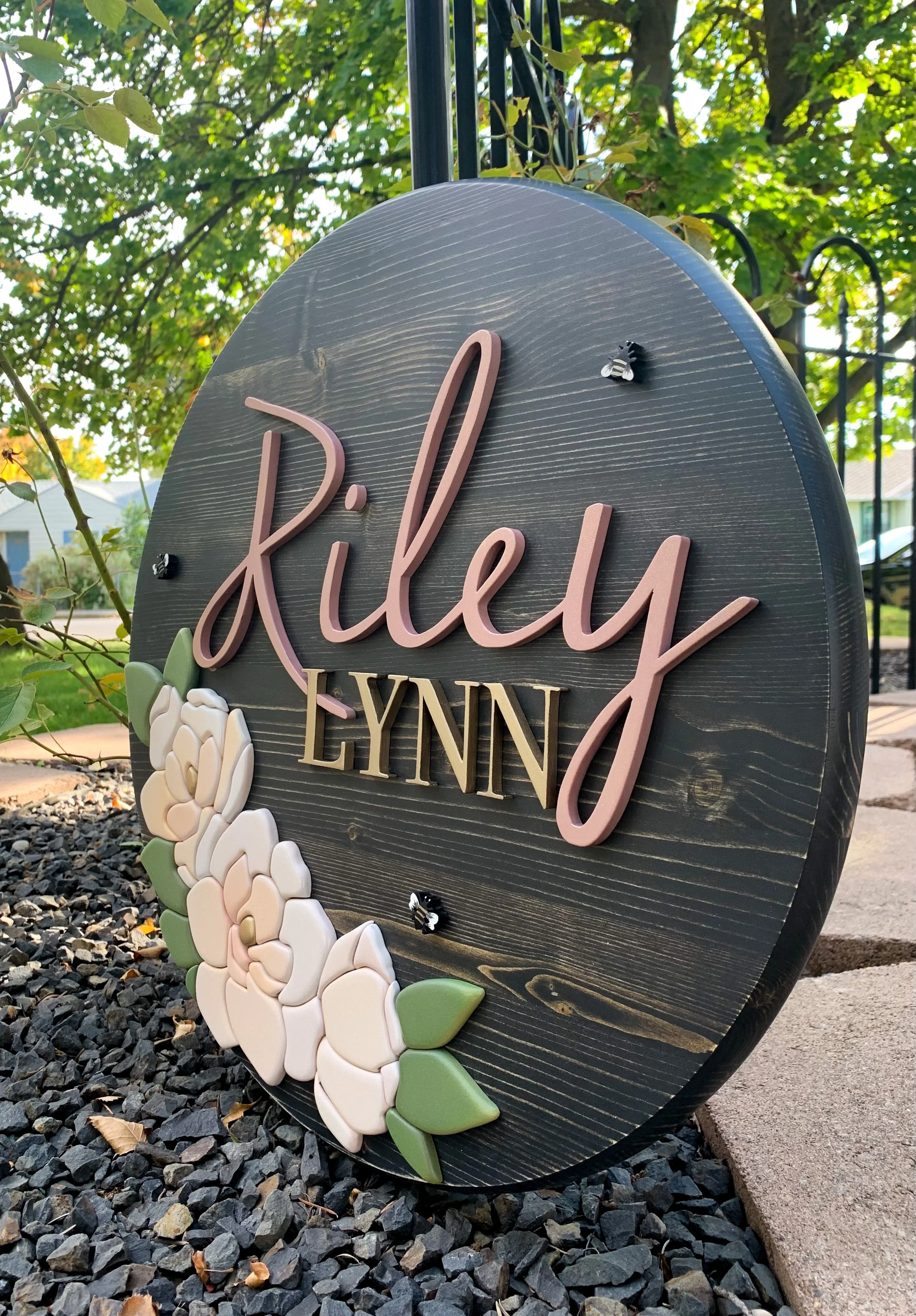 Decorative kids room sign on a dark wooden background, decorated with white and pink floral accents and small bee embellishments.