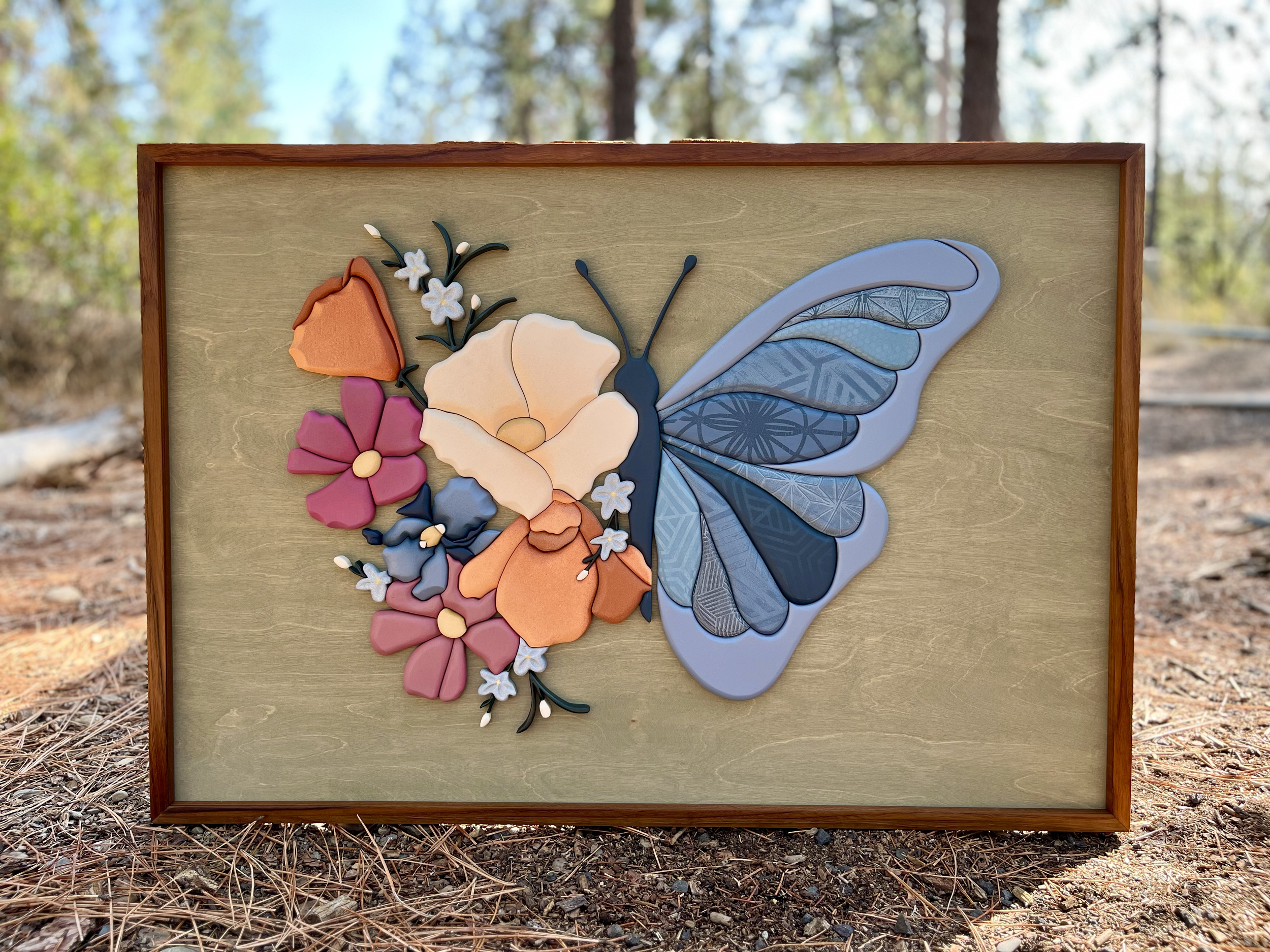 A framed artwork of a butterfly with colorful geometric flower designs on a wooden background, placed outdoors on a ground covered with pine needles, with trees and sky in the background.