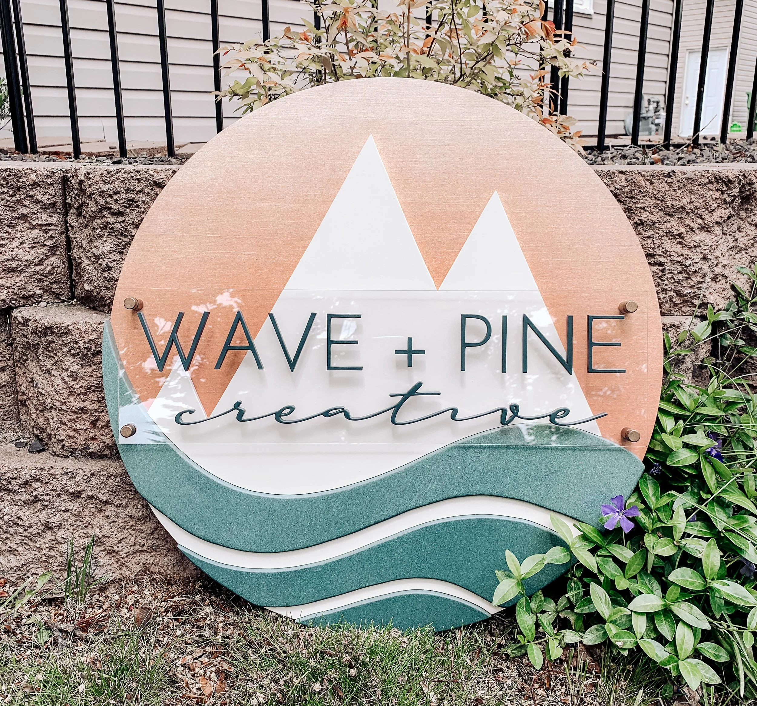 A decorative sign reading "WAVE + PINE creating" in front of a brick wall with plants and grass around it.