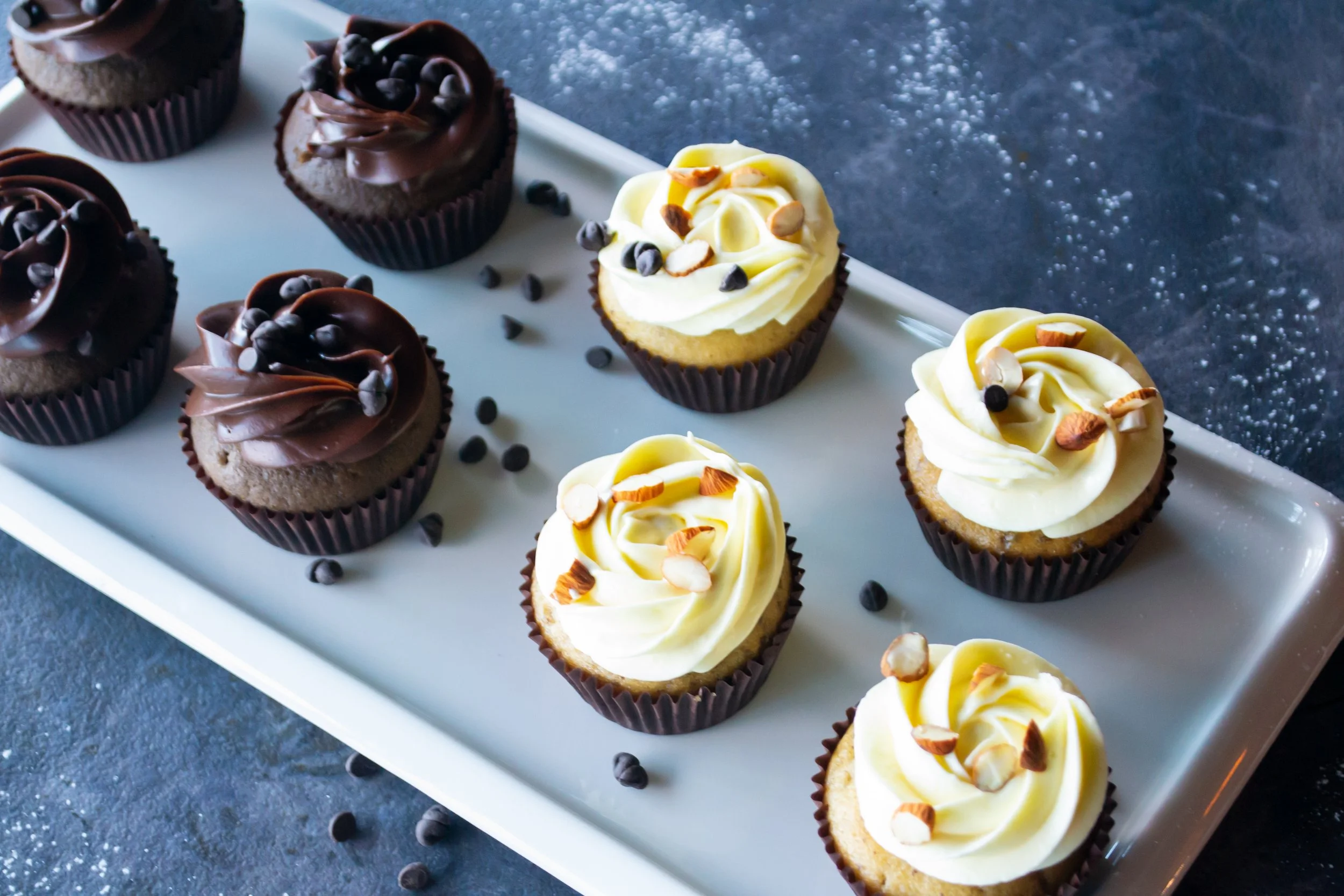 Assorted cupcakes with chocolate and vanilla frosting topped with chocolate chips and chopped nuts, arranged on a white serving platter with scattered chocolate chips nearby.