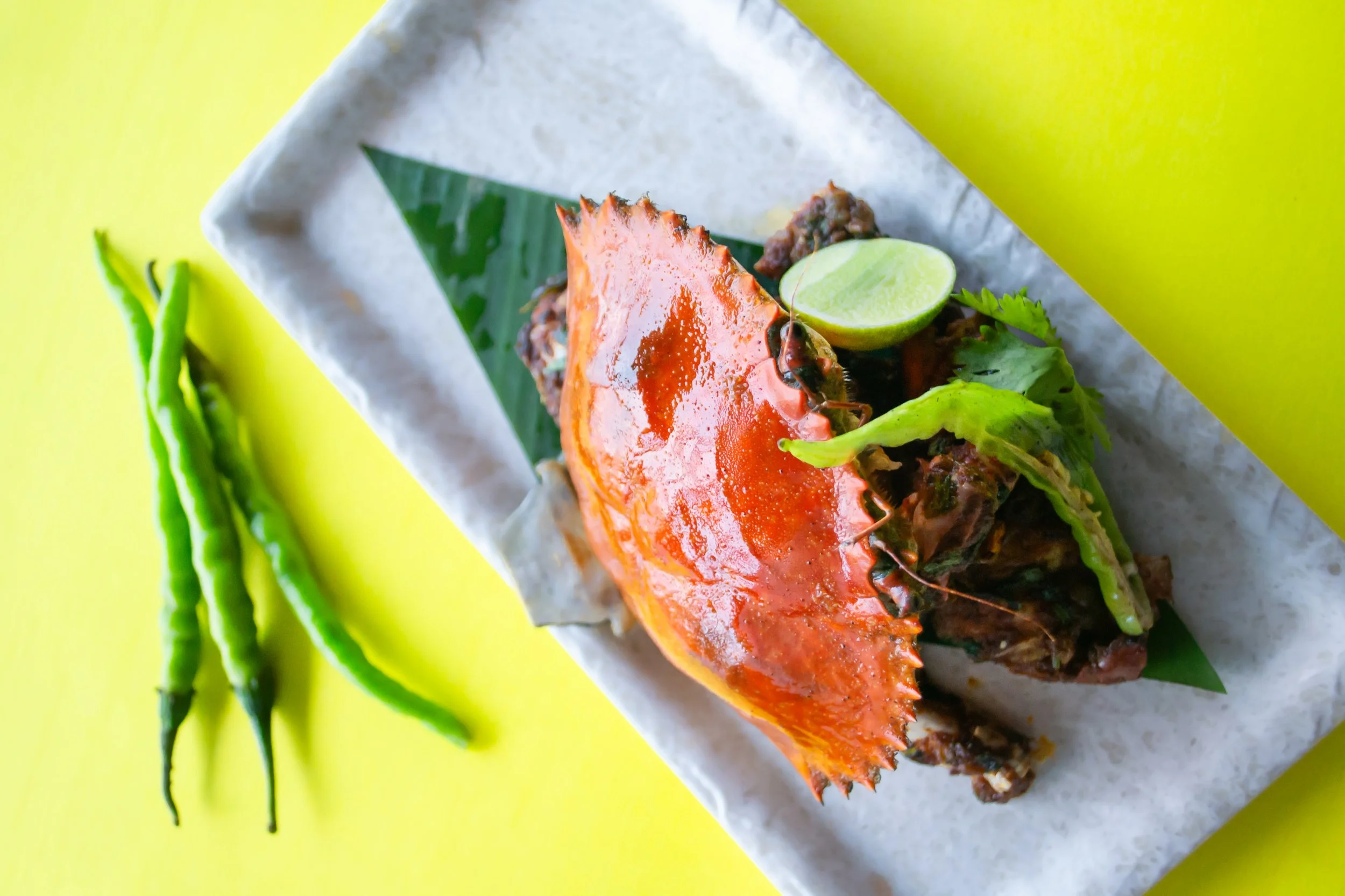 Cooked crab with lime wedge, green chili, and cilantro on a white paper-lined yellow surface.