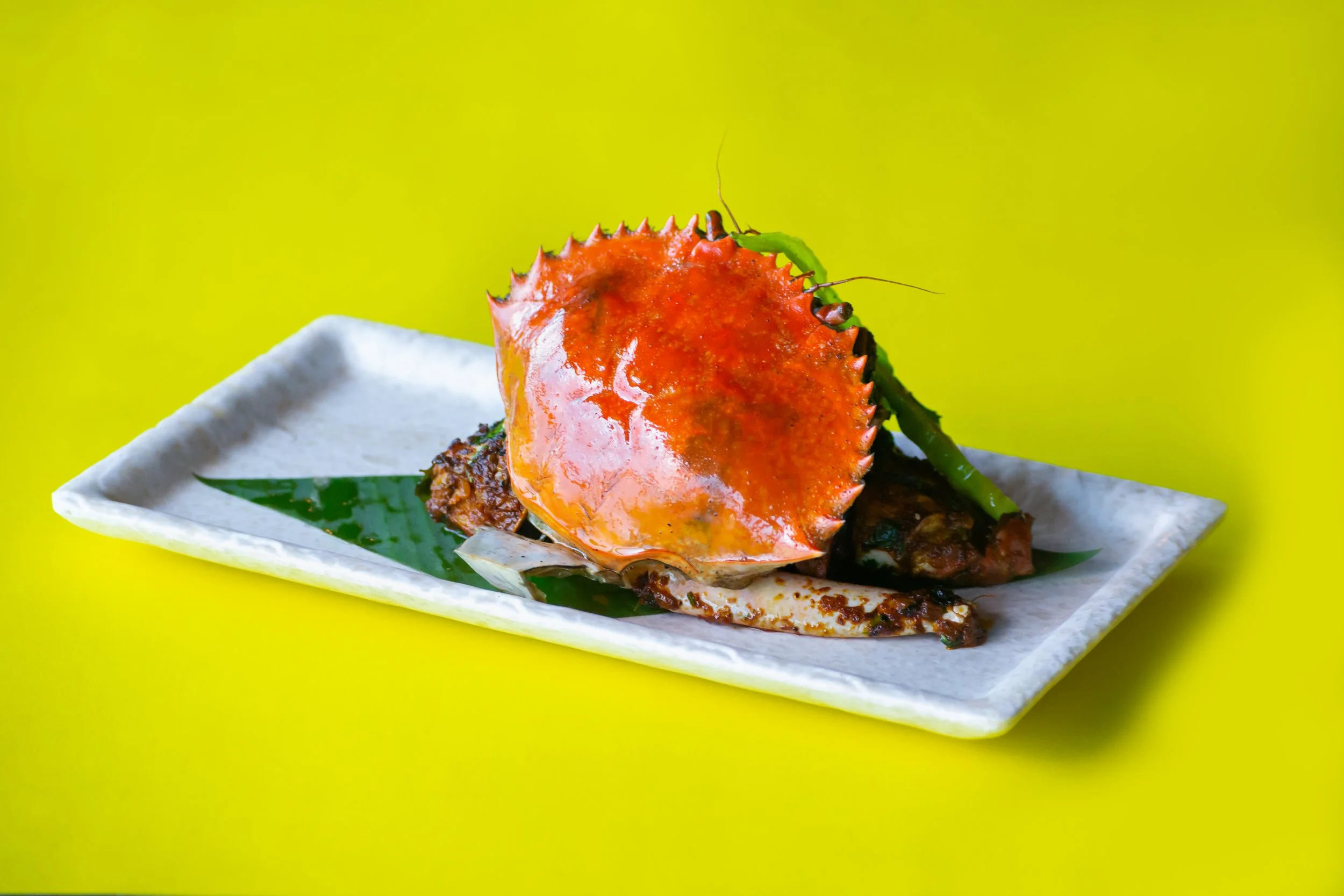 A cooked crab with an orange shell served on a white rectangular dish with a green leaf underneath, against a yellow background.